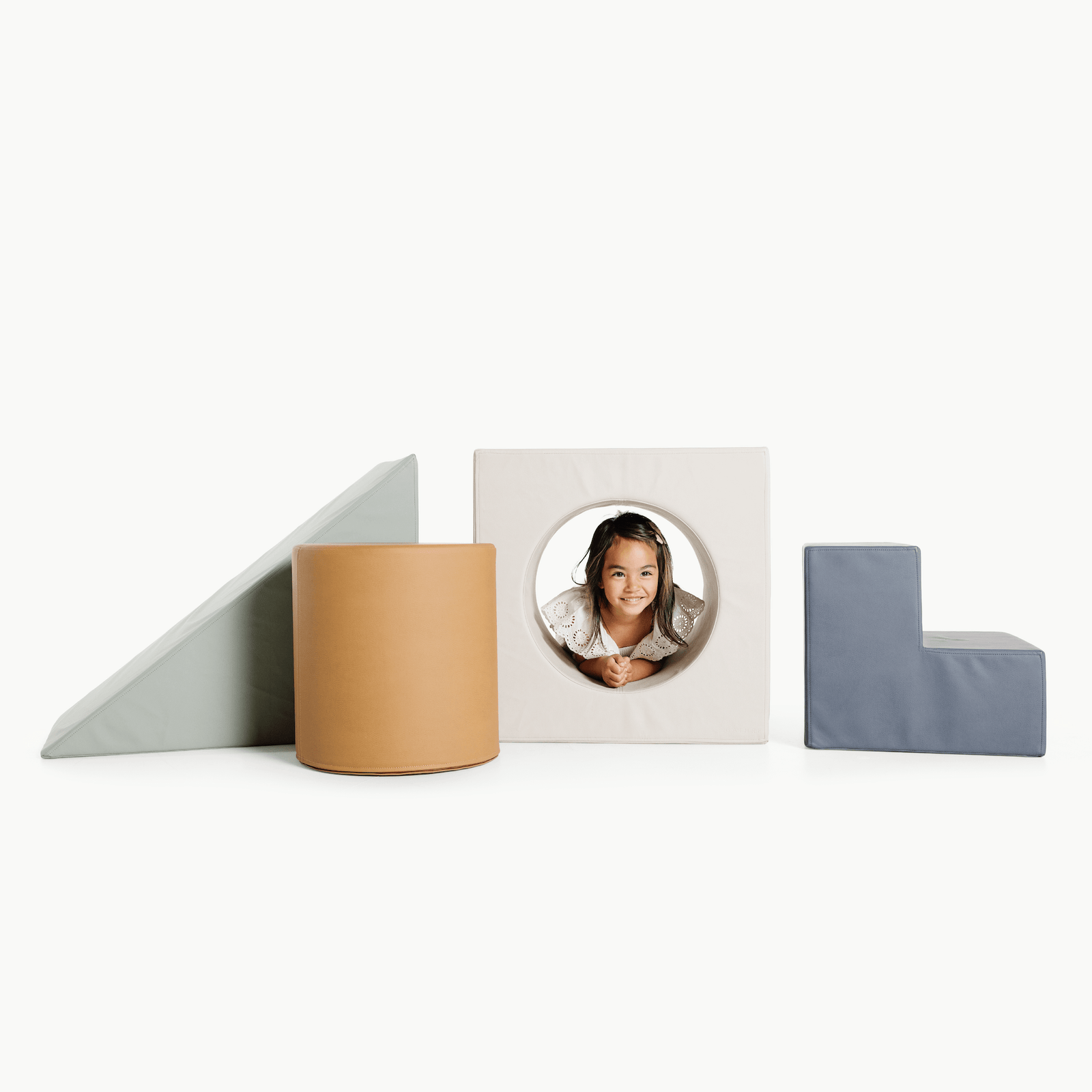 A child smiles while peeking through the Wholesale Block Playset by Gathre, surrounded by soft white foam shapes—a wedge, a cylinder, and an L-shaped seat—against a white background.
