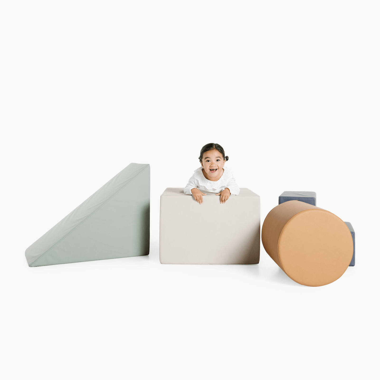 A smiling child peeks over a large, light-colored foam block from the Gathre Wholesale Block Playset, surrounded by soft geometric shapes—wedge, cylinder, and cubes—on a white background.