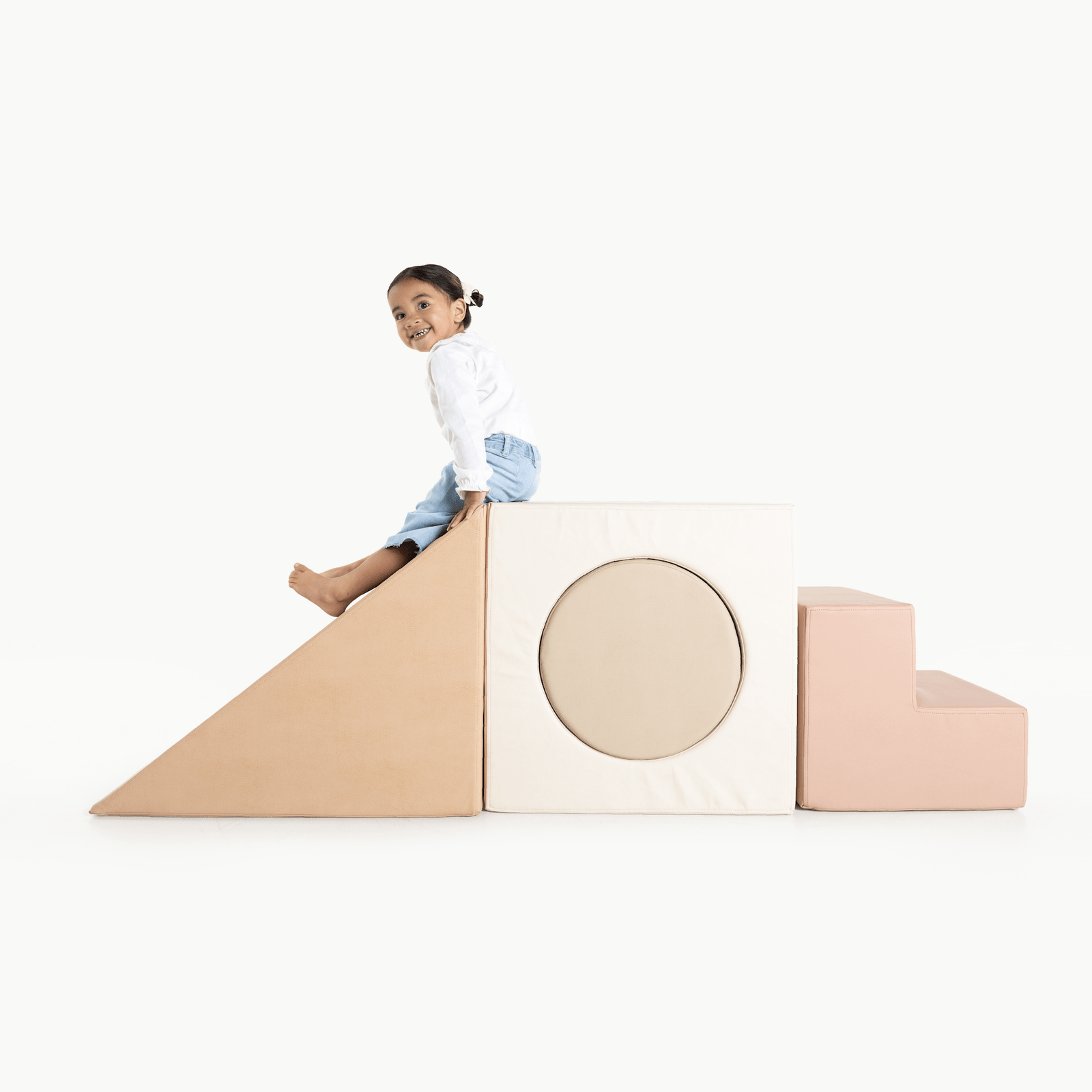 A young child smiles while sitting atop the Gathre Wholesale Block Playset, featuring soft, pastel-colored foam blocks—including steps, a square with a circular hole, and a ramp—against a white background.
