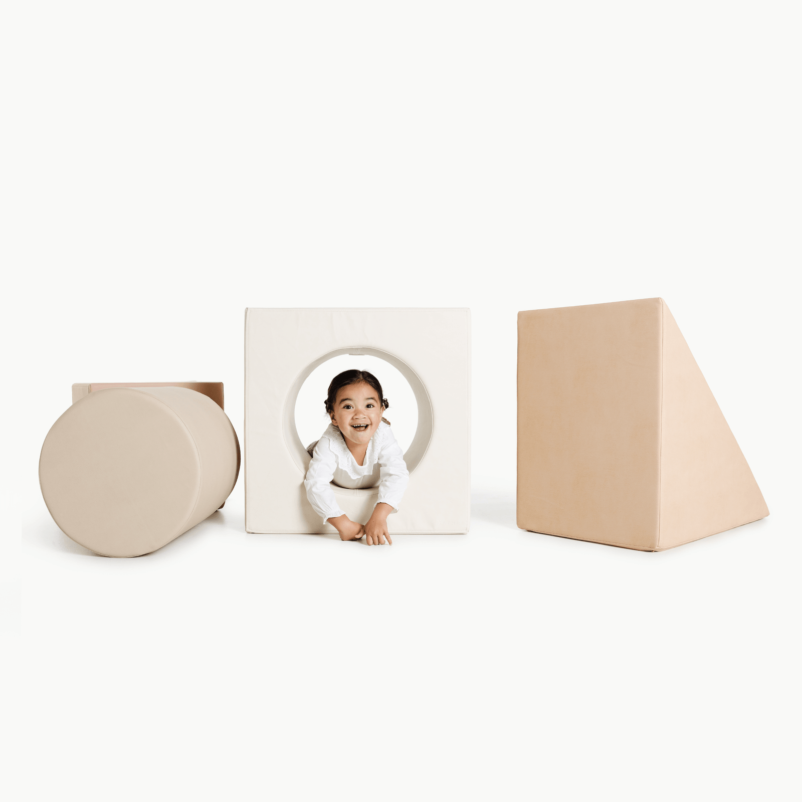 A smiling young child crawls through a circular opening in the soft, white Gathre Wholesale Block Playset, surrounded by large beige foam shapes on a white background.
