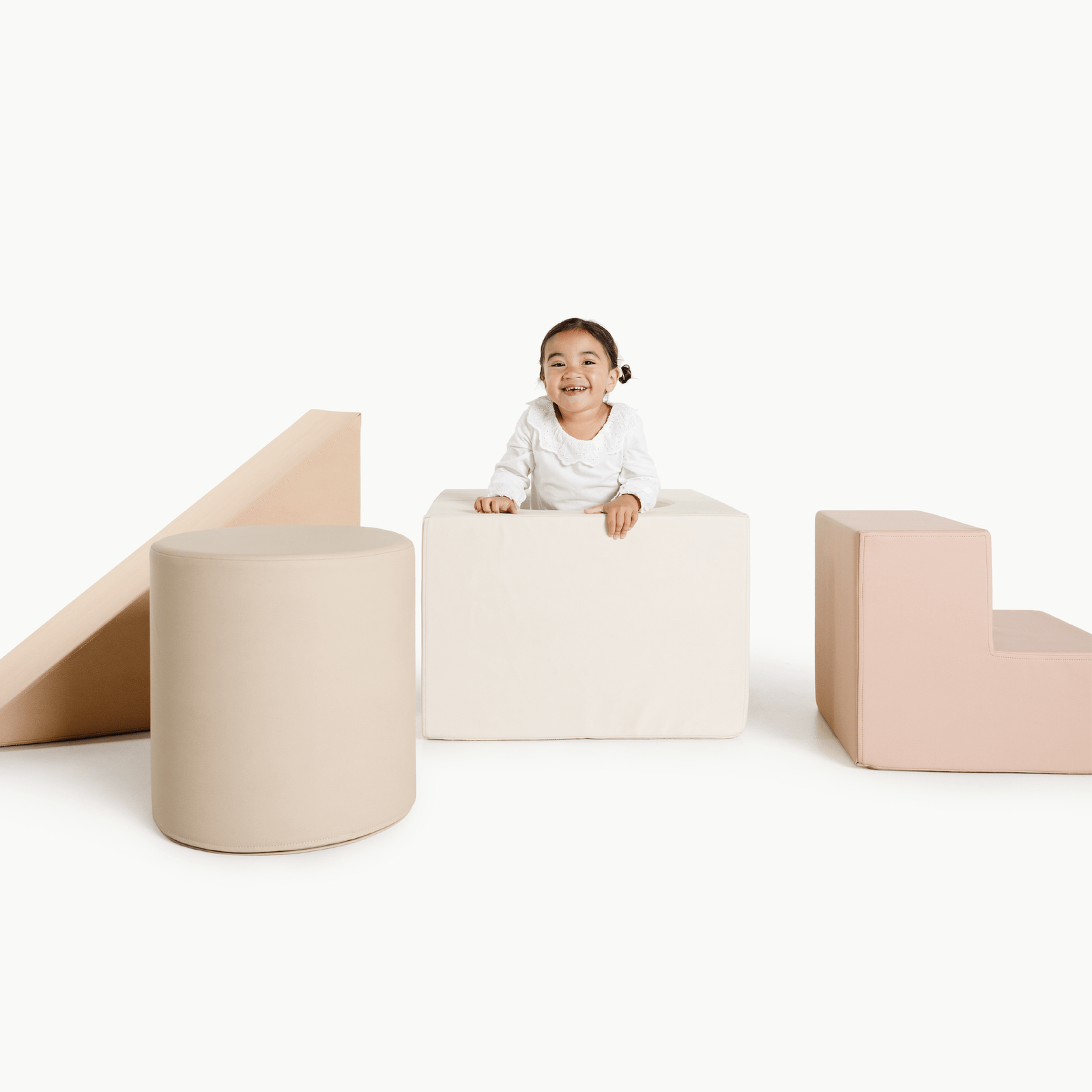 A smiling child sits on a large white foam block from the Gathre Wholesale Block Playset, surrounded by beige and pink foam shapes—cylinder, ramp, and L-block—against a plain white background.