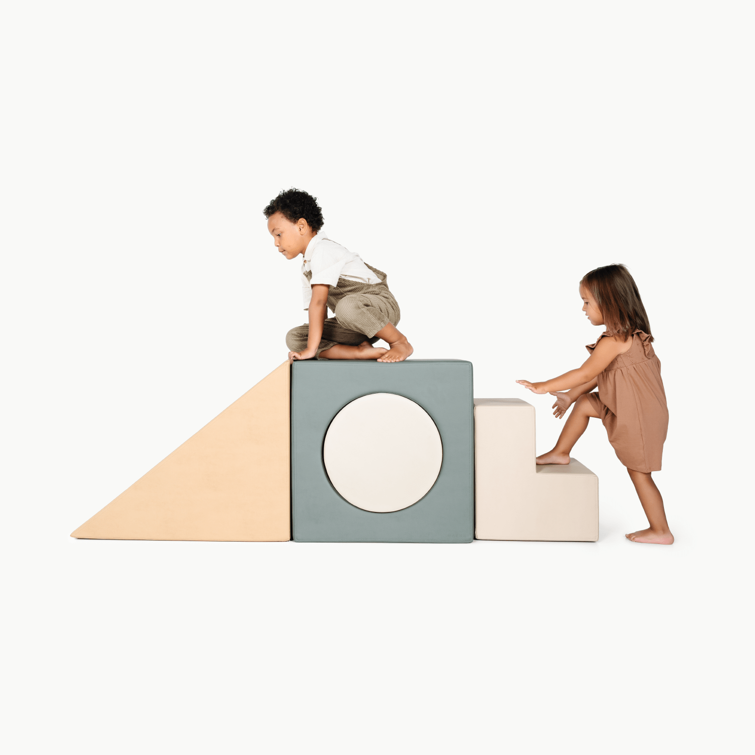 Two young children play on the Gathre Wholesale Block Playset. One sits atop a gray cube, while the other climbs beige steps, both set against a white background.