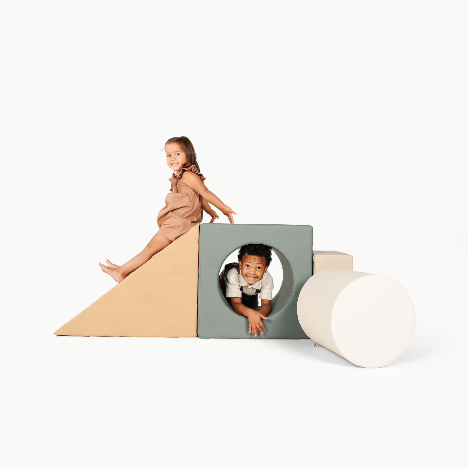 Two smiling children play with the Gathre Wholesale Block Playset. A girl sits on a tan ramp, while a boy crawls through a green block with a circular hole, both looking happy and playful.