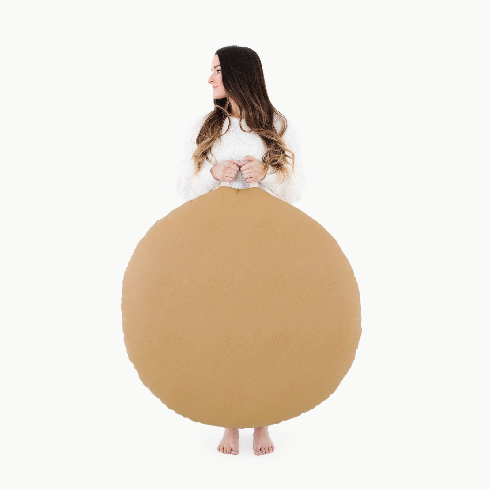 A woman with long hair stands barefoot against a white background, holding a large, round Gathre Wholesale Floor Cushion in Camel that covers her body from the chest down as she looks to her left.
