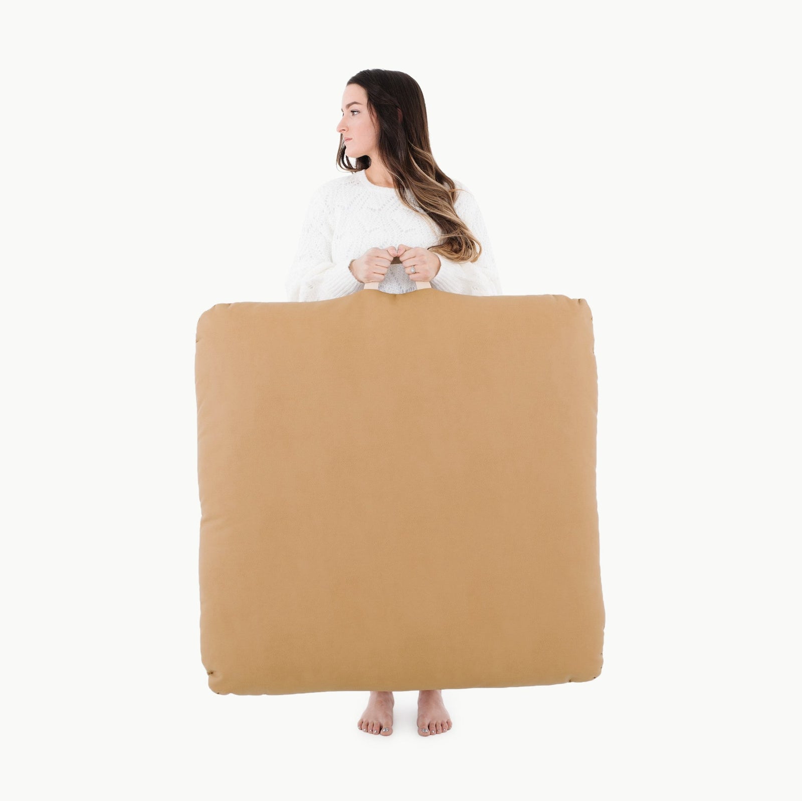 A woman in a white sweater stands barefoot, holding a large, square Gathre Wholesale Floor Cushion in Camel from shoulders to ankles, looking left against a plain white background.