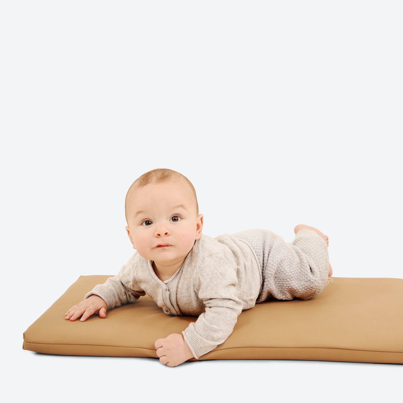 A baby in a light outfit lies on their stomach on the Gathre Wholesale Padded Changing Mat, gazing up with a neutral expression against a plain white background.