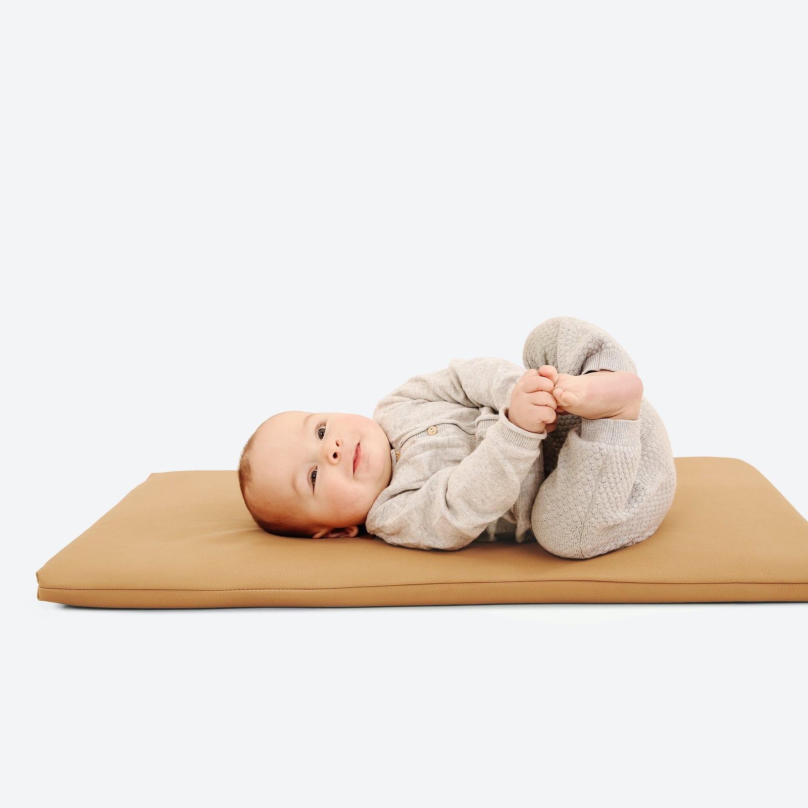A baby in a gray outfit lies on the Gathre Wholesale Padded Changing Mat in Camel, holding their feet and gazing upwards against a plain white background.