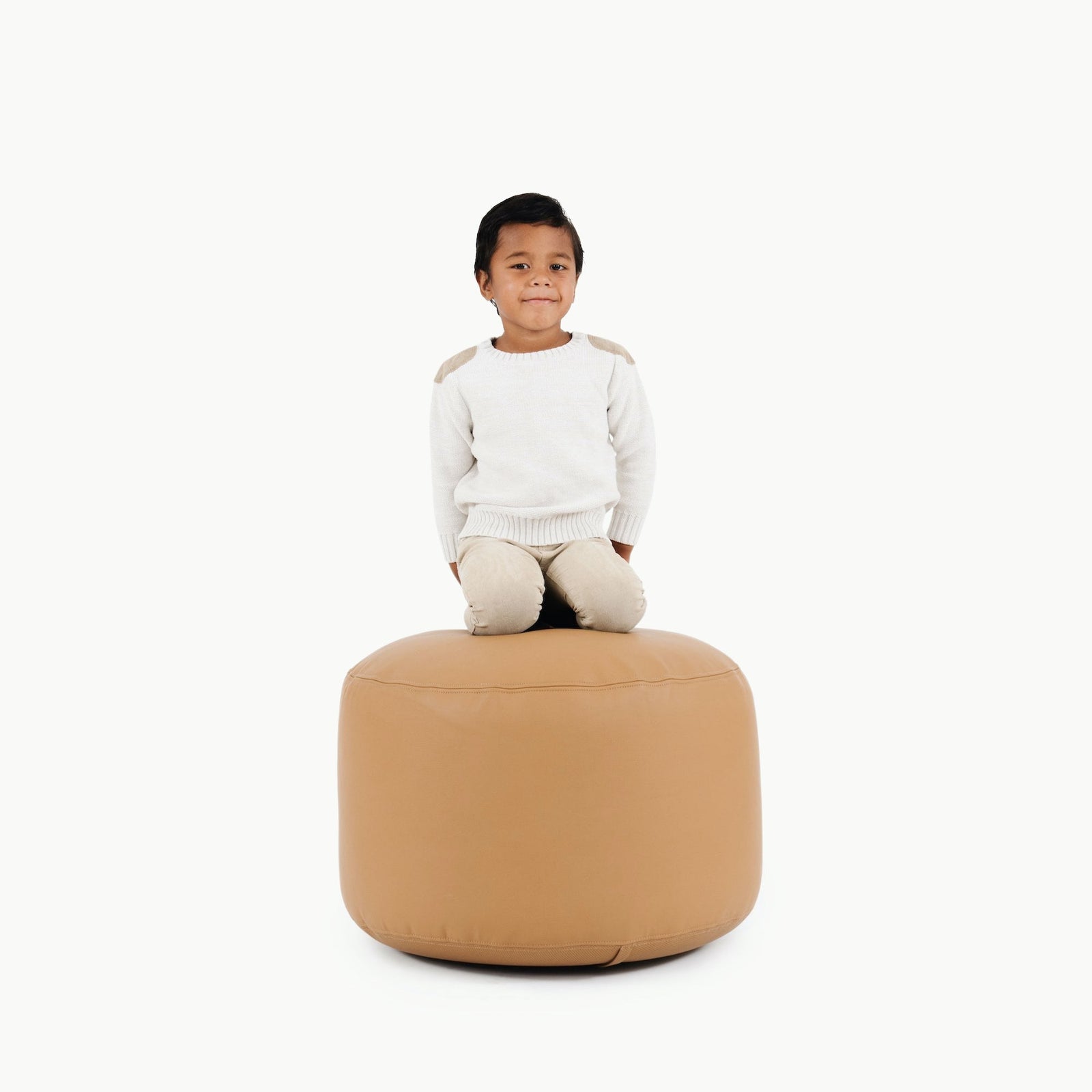 A young child in a white sweater and beige pants kneels on the Gathre Wholesale Pouf in Camel, set against a plain white background.
