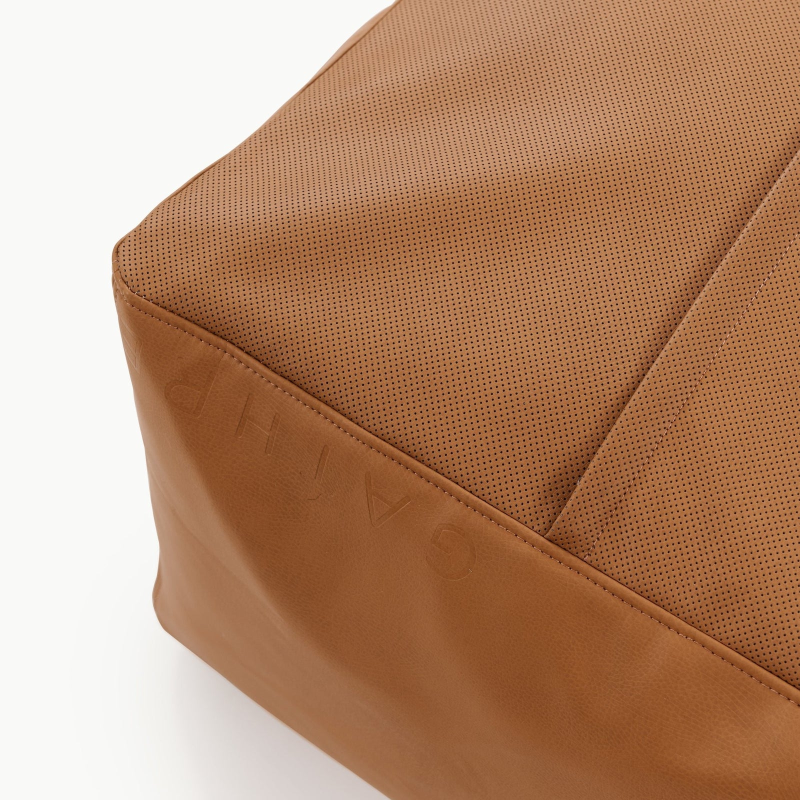 Close-up of the corner of a tan-colored, perforated leather Wholesale Pouf by Gathre, with embossed letters partially visible on the side, set against a plain white background.