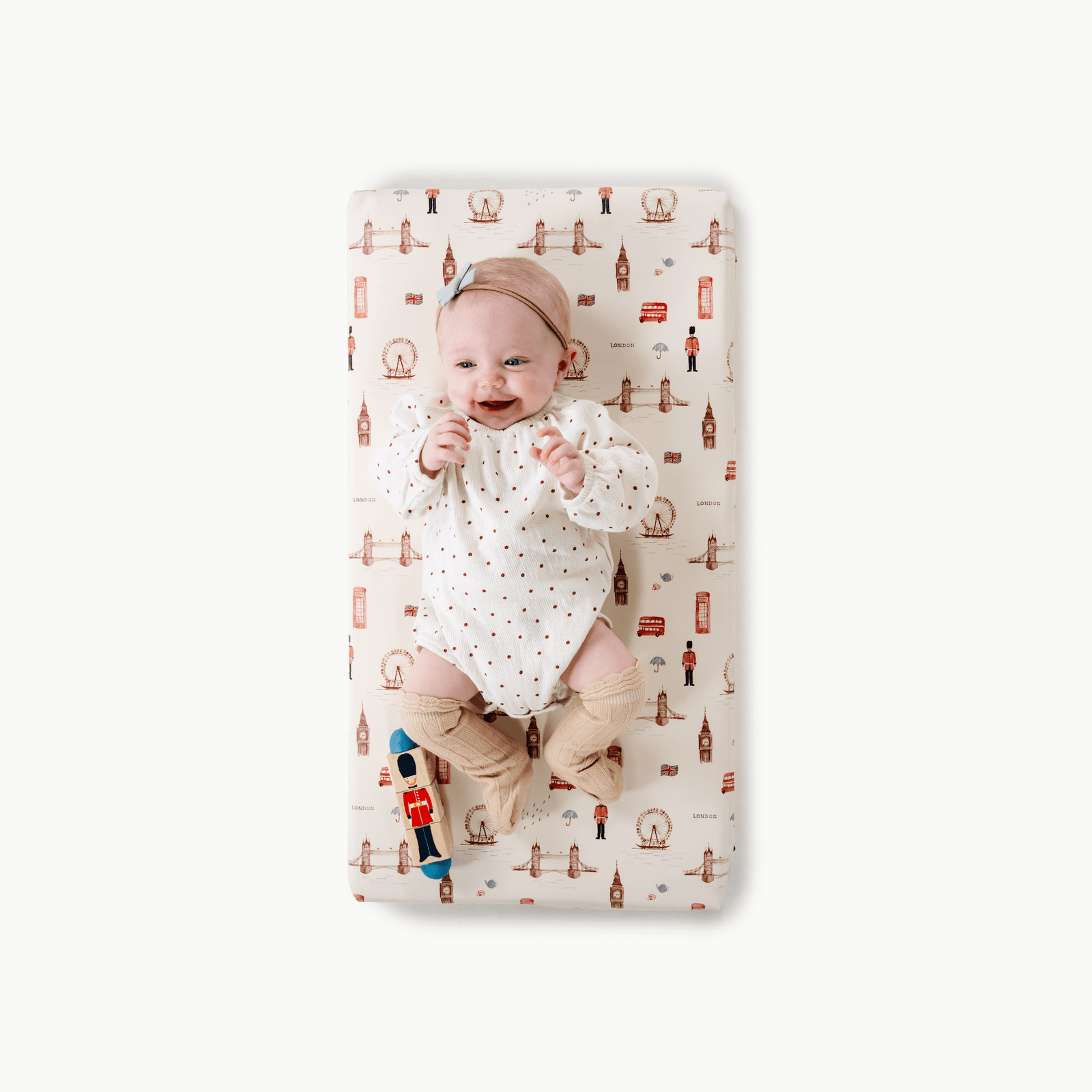 A smiling baby in a white polka-dot onesie and beige booties lies on the Gathre Wholesale Padded Changing Mat featuring London-themed illustrations, with a red and blue toy by the baby's left foot.