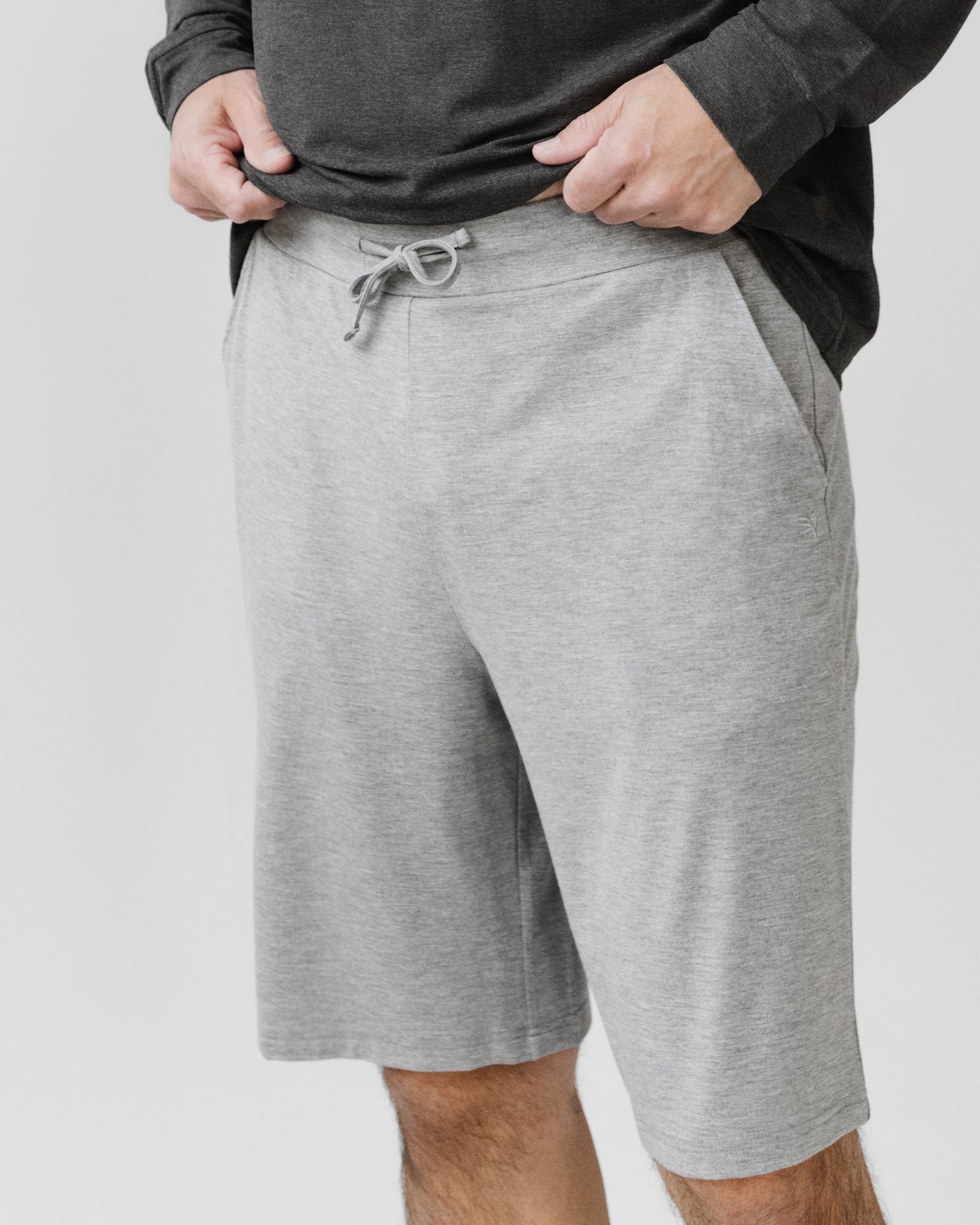 A person wears Cozy Earth's Men's Bamboo Stretch-Knit Short Sleeve Pajama Set in light gray, hands in pockets. The background is plain and light-colored.