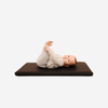 A baby lies on their back on a Gathre Wholesale Padded Changing Mat in Raven, holding their feet with both hands and looking up against a white background.