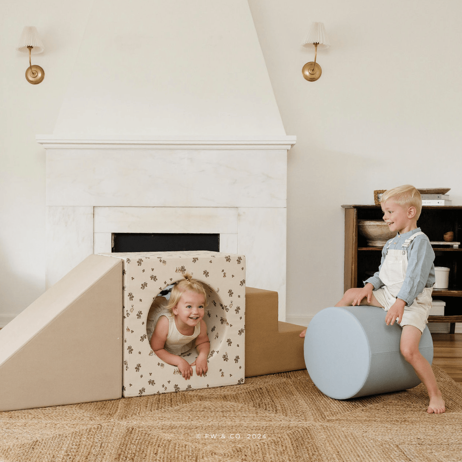 Two children enjoy the Gathre Wholesale Block Playset indoors; one crawls through a printed cube with a circular hole, while the other sits on a large cylinder block. Both kids smile in a cozy, well-lit room.