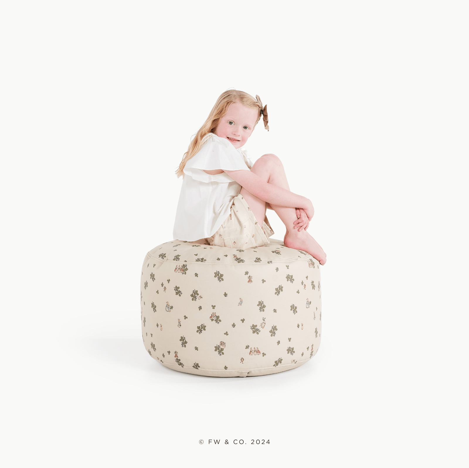 A young child with long blonde hair and a clip sits on a Gathre Wholesale Pouf in Peter Rabbit™ color, wearing a white top and beige skirt, against a plain white background.