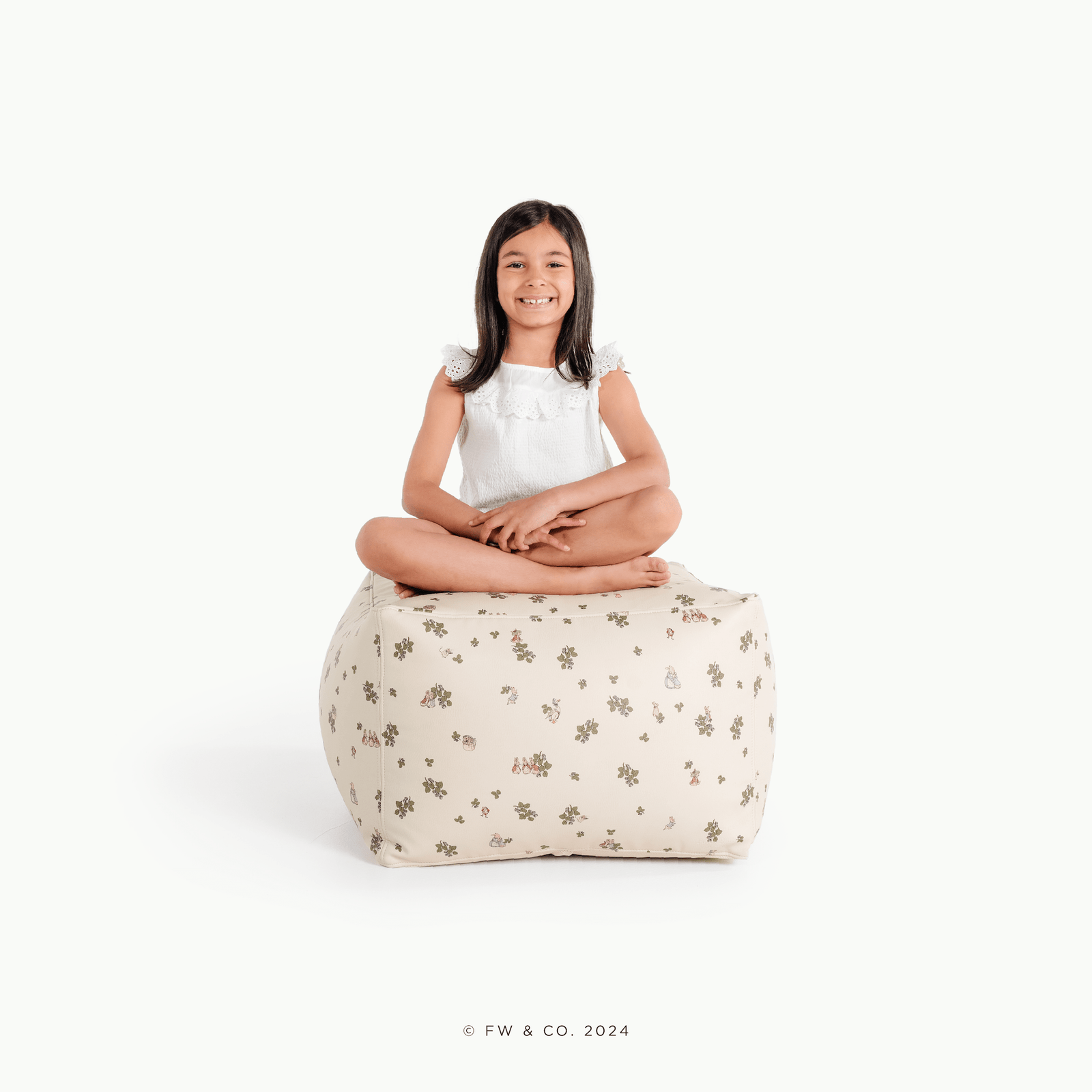 A smiling young girl in a white dress sits cross-legged on the Gathre Wholesale Pouf in Peter Rabbit™ color, set against a plain white background.