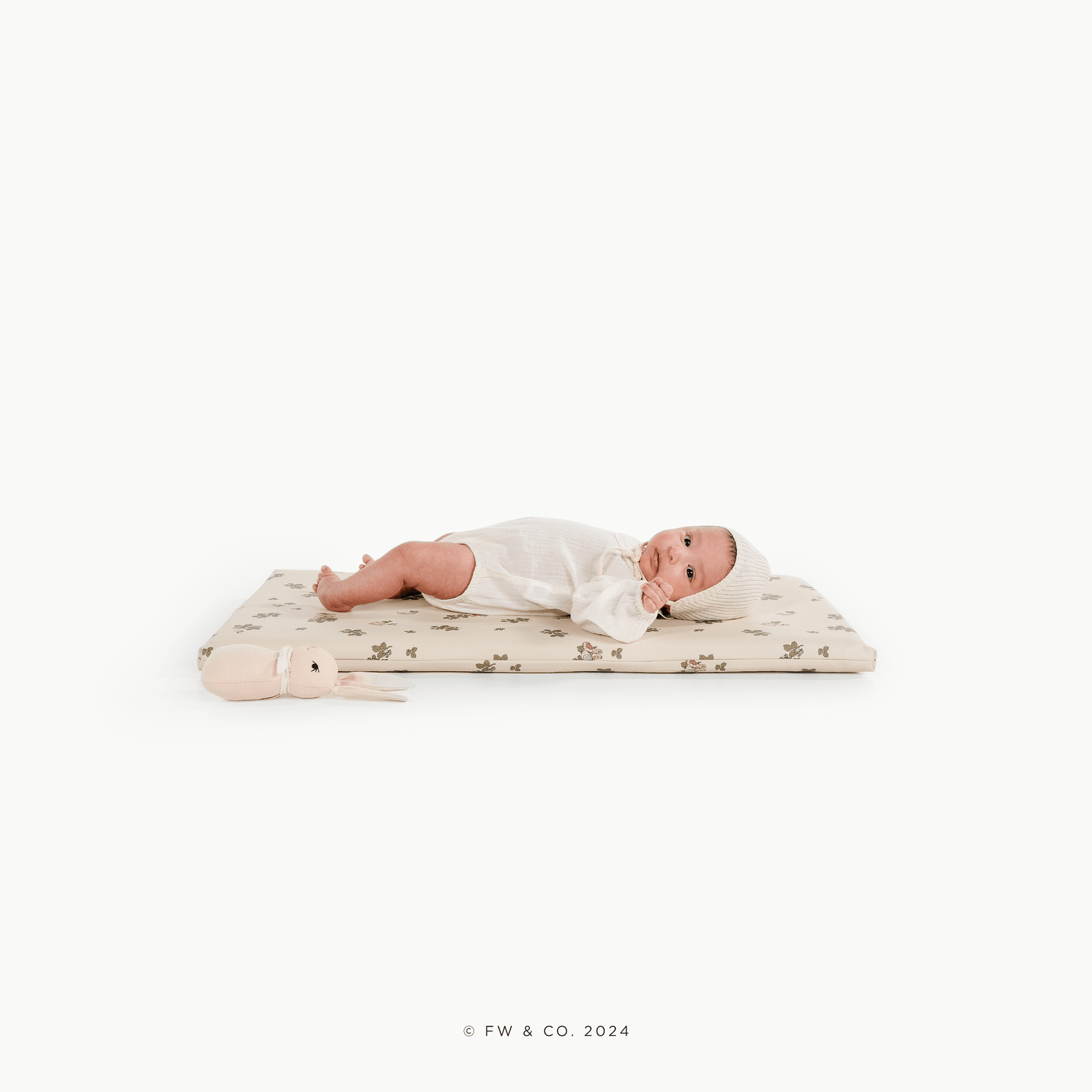 A baby rests on the Gathre Wholesale Padded Changing Mat in Peter Rabbit™ color, next to a soft bunny toy, all set against a plain white background.
