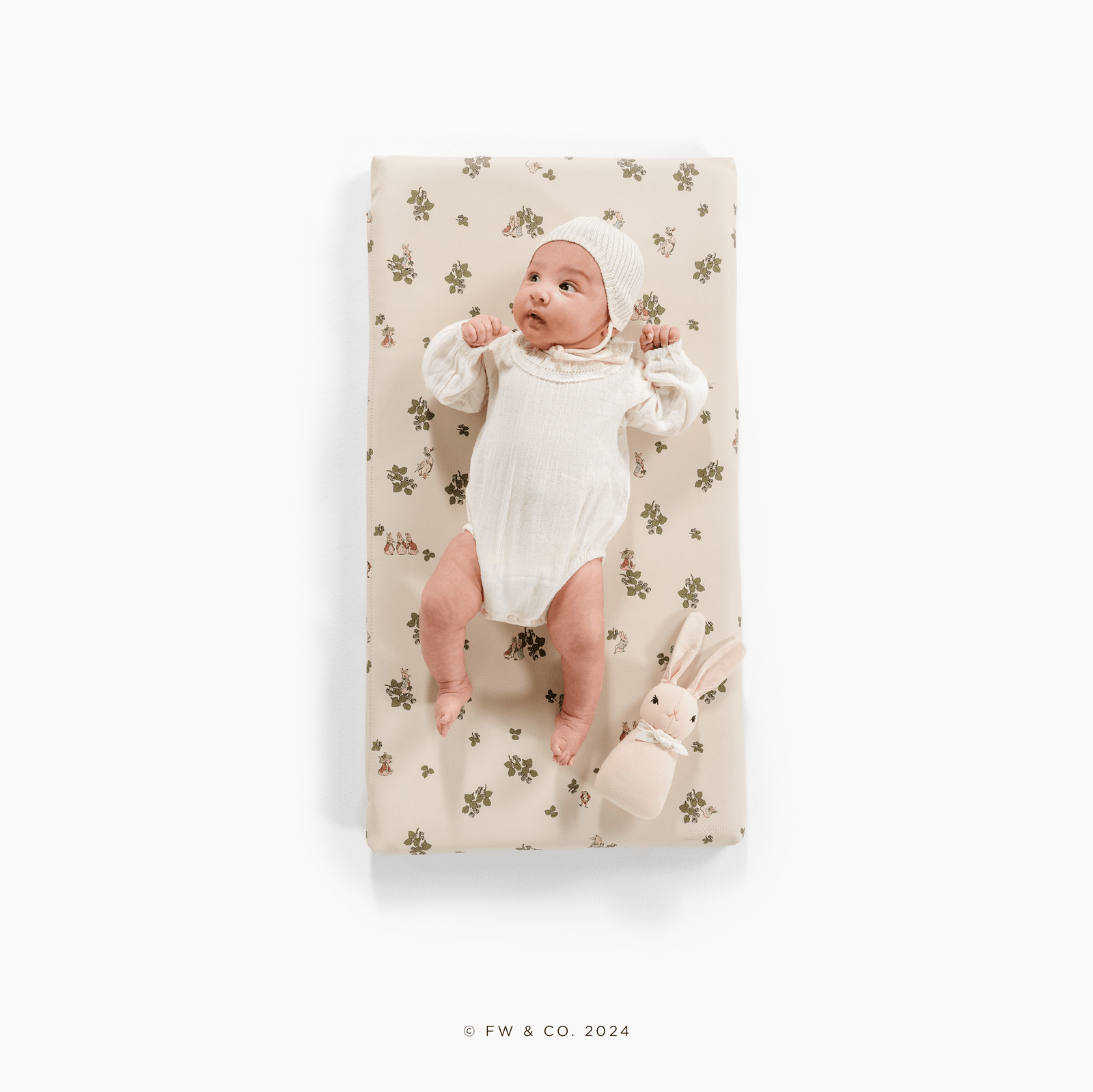A baby in a white outfit and knit bonnet lies on the Gathre Wholesale Padded Changing Mat with a floral pattern, next to a soft beige bunny toy, all set against a white background.