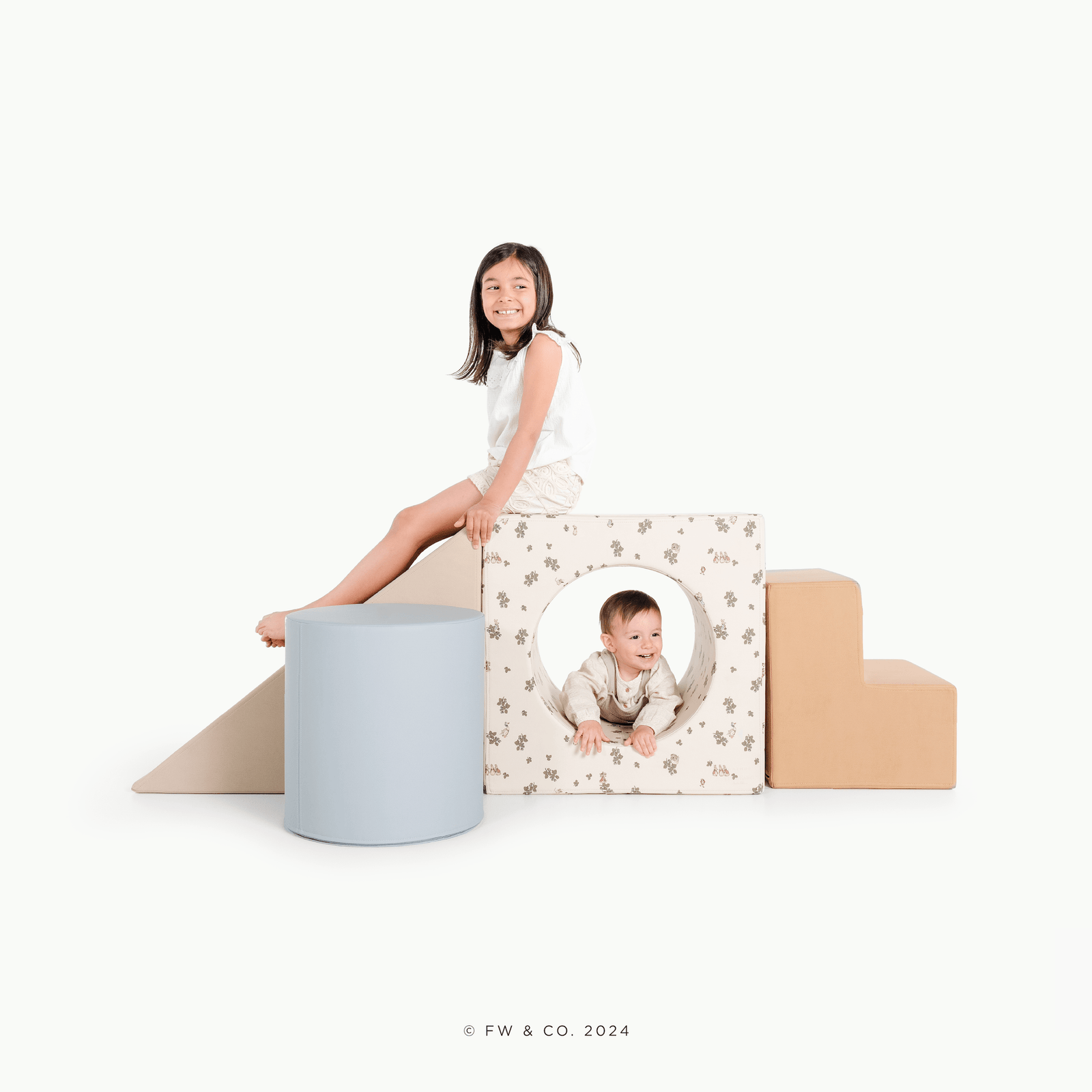 A smiling young girl sits on a beige staircase from the Gathre Wholesale Block Playset, next to a blue cylinder, while a baby peeks out from a floral-patterned cube with a circular cutout. The background is white.