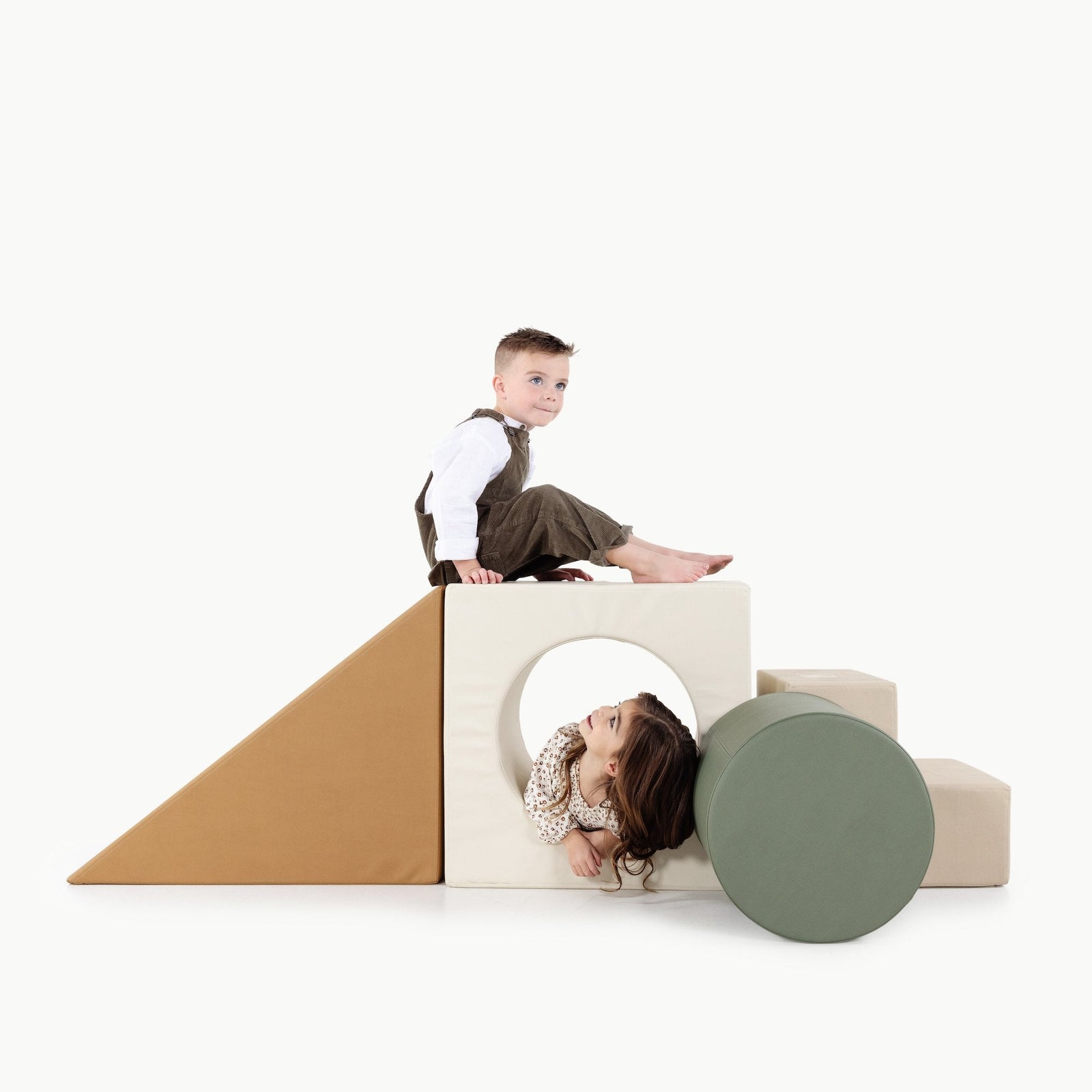 Two children play with the Gathre Wholesale Block Playset. One sits atop a cube with a circular hole while the other crawls through it. The set features soft geometric shapes, including a ramp, steps, and a large green cylinder.