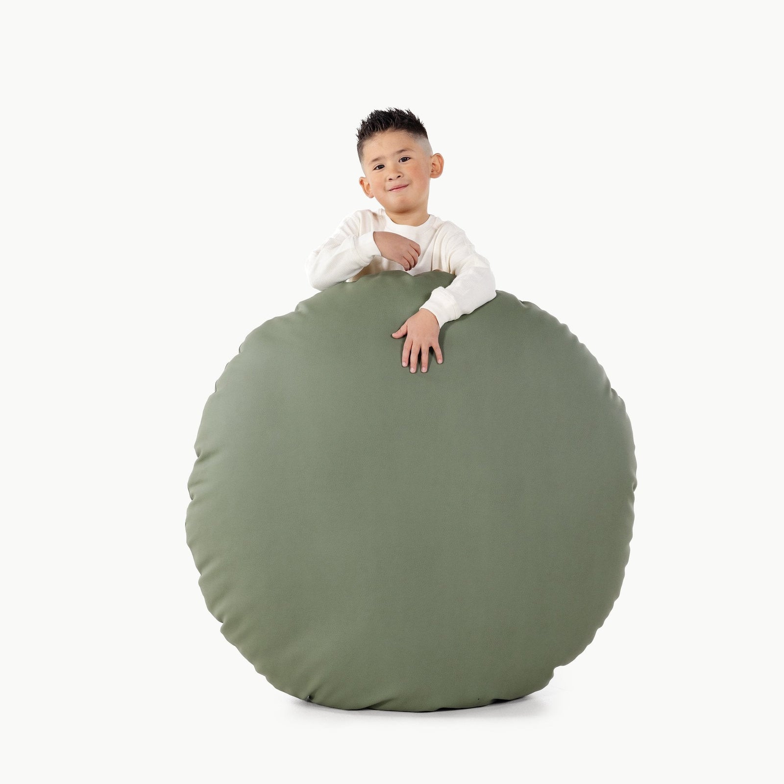 A young boy with short dark hair and a white long-sleeve shirt rests his arms on a large, round, sage-green Gathre Wholesale Floor Cushion against a plain white background.