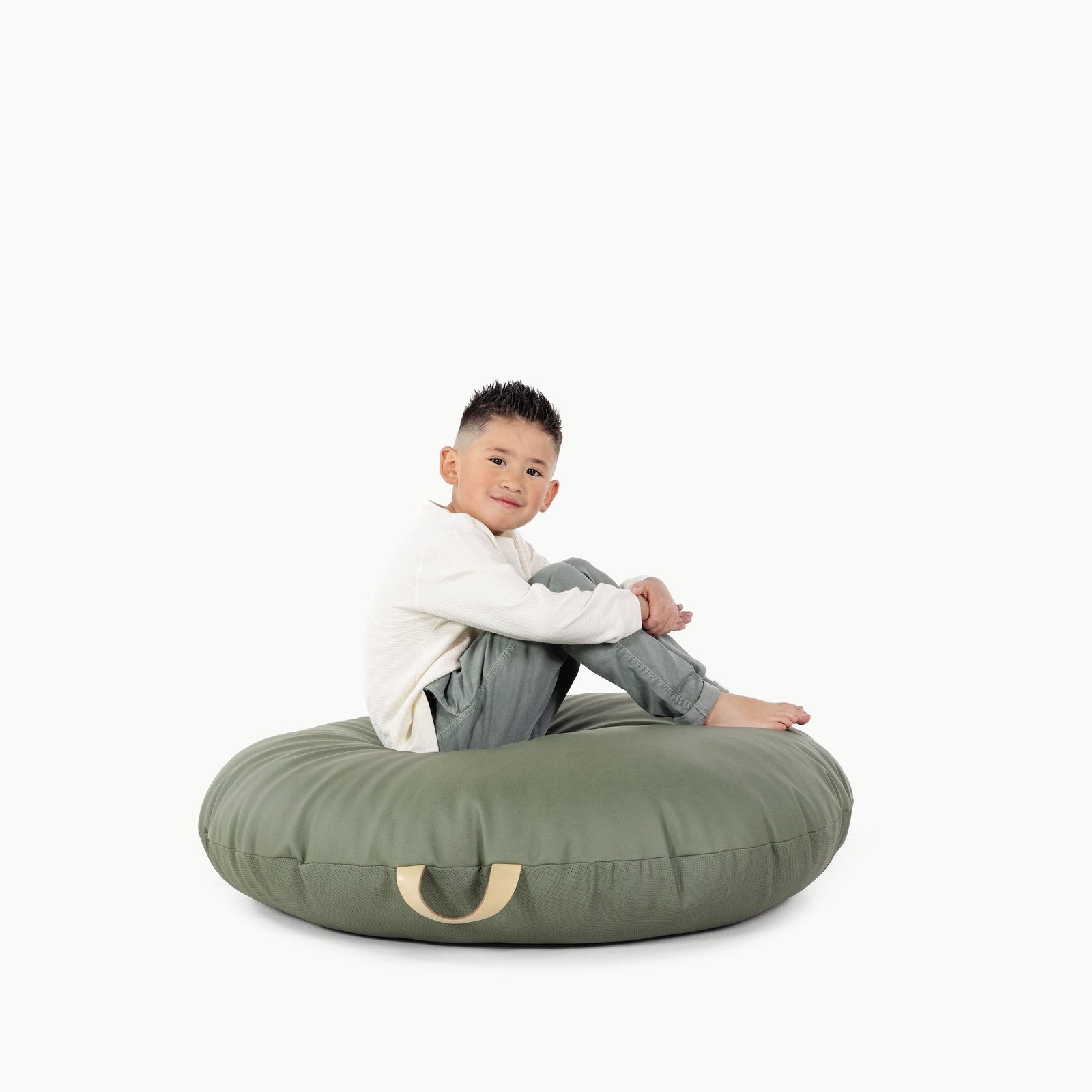 A young boy sits barefoot with his knees up on the Gathre Wholesale Floor Cushion in Thyme, featuring a carrying handle, against a plain white background.