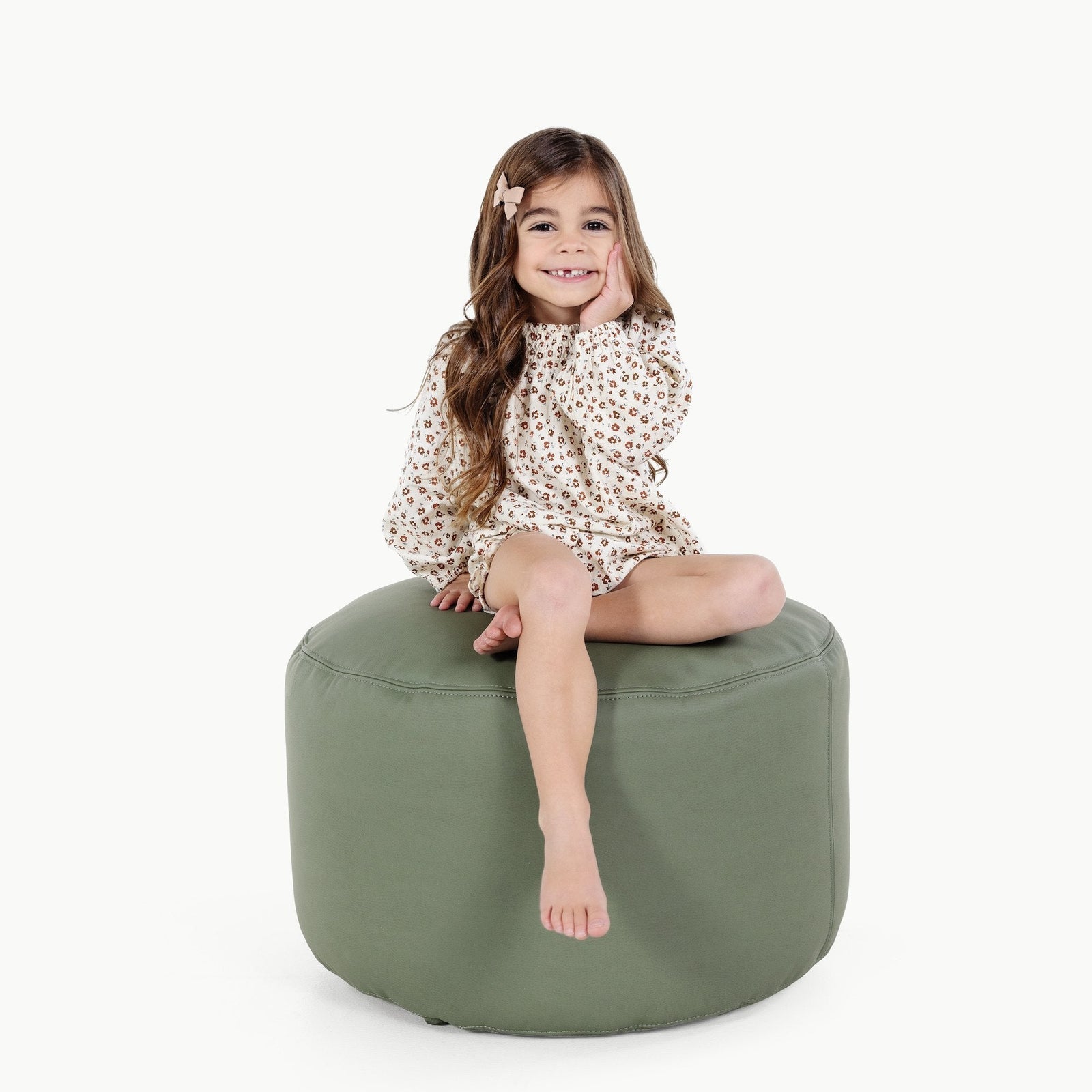 A young girl with long brown hair and a floral dress sits barefoot, smiling with one hand on her cheek, on the Gathre Wholesale Pouf in Thyme against a white background.
