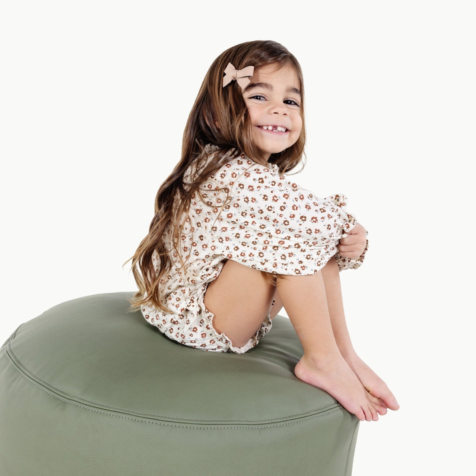 A young girl with long brown hair and a bow clip smiles while sitting cross-legged on a Gathre Wholesale Pouf in olive green, wearing a cream floral outfit against a plain white background.