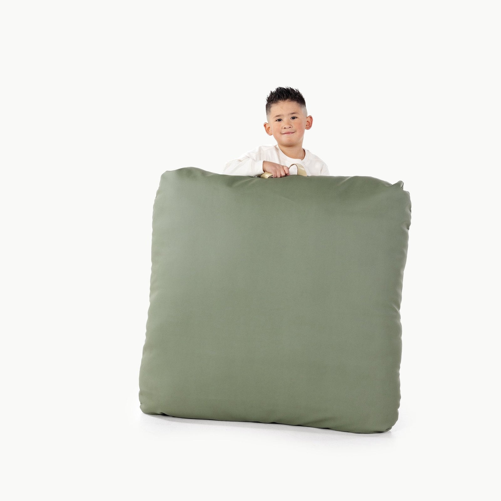 A young boy peeks over a large, square Wholesale Floor Cushion by Gathre, featuring a handle, set against a plain white background.