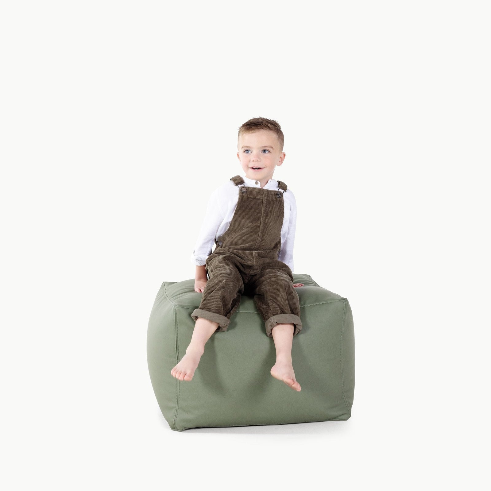 A young boy in brown overalls and a white shirt sits barefoot on a Thyme Wholesale Pouf by Gathre against a plain white background.