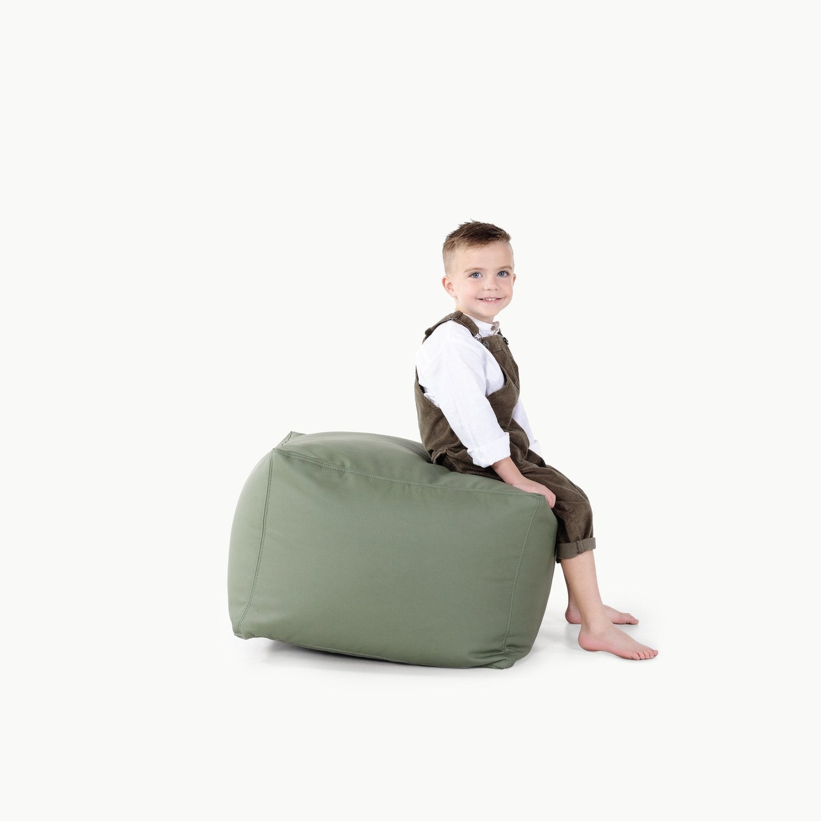 A young boy with short hair, wearing a white shirt and brown overalls, sits barefoot on the green Gathre Wholesale Pouf against a plain white background.