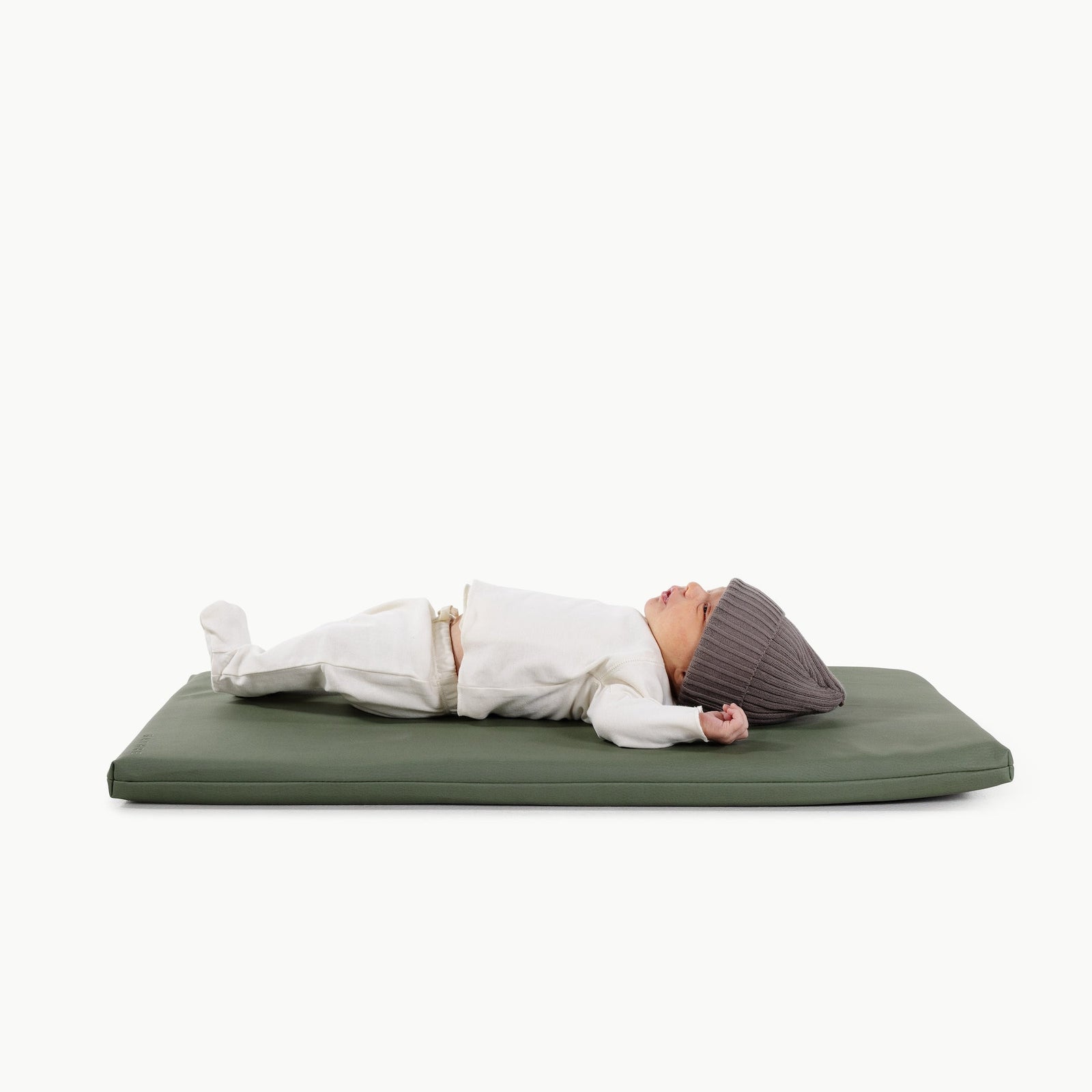 A newborn baby wearing a gray knit hat and white outfit lies on a Gathre Wholesale Padded Changing Mat in Thyme color, set against a plain white background.