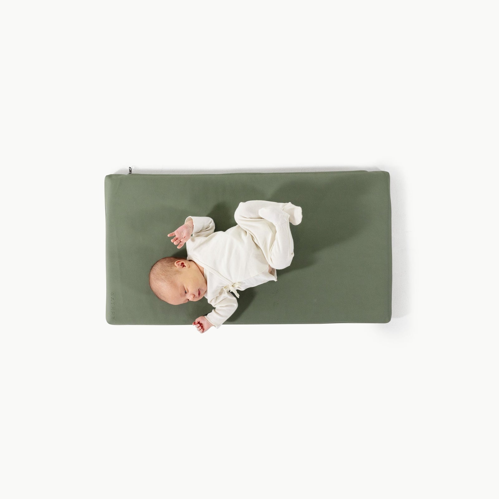 A newborn lies on its back, arms and legs gently raised, on a rectangular Wholesale Padded Changing Mat by Gathre in olive green. The baby wears a white outfit, positioned slightly left against a plain white background.