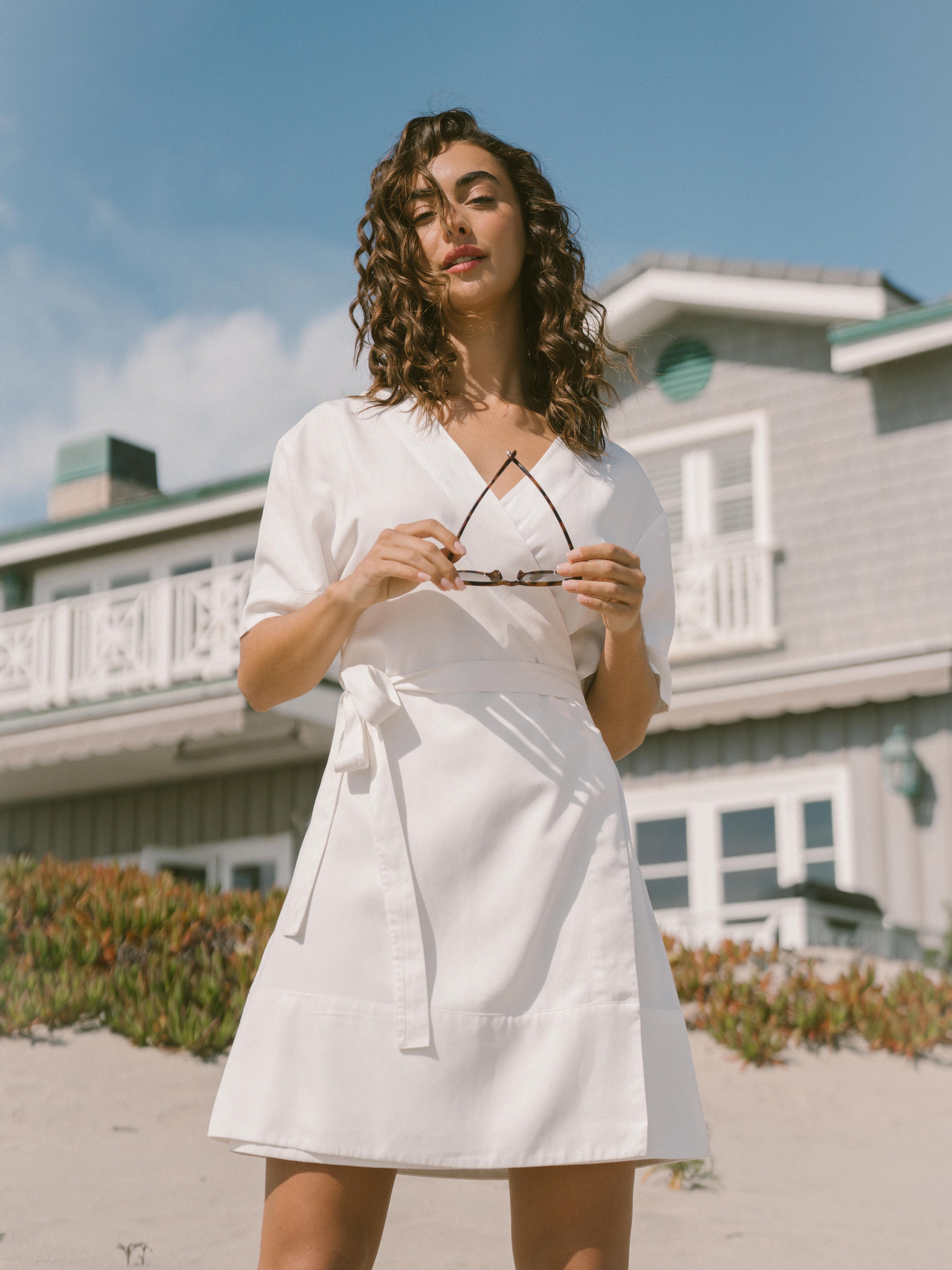 A woman with curly hair is standing in front of a house. She is wearing a White Women's Cozy Earth Coastal Comfort Wrap Dress by Cozy Earth and holding a pair of sunglasses in her hands. The background features a two-story house with a porch and railing, set against a partly cloudy sky. |Color:White