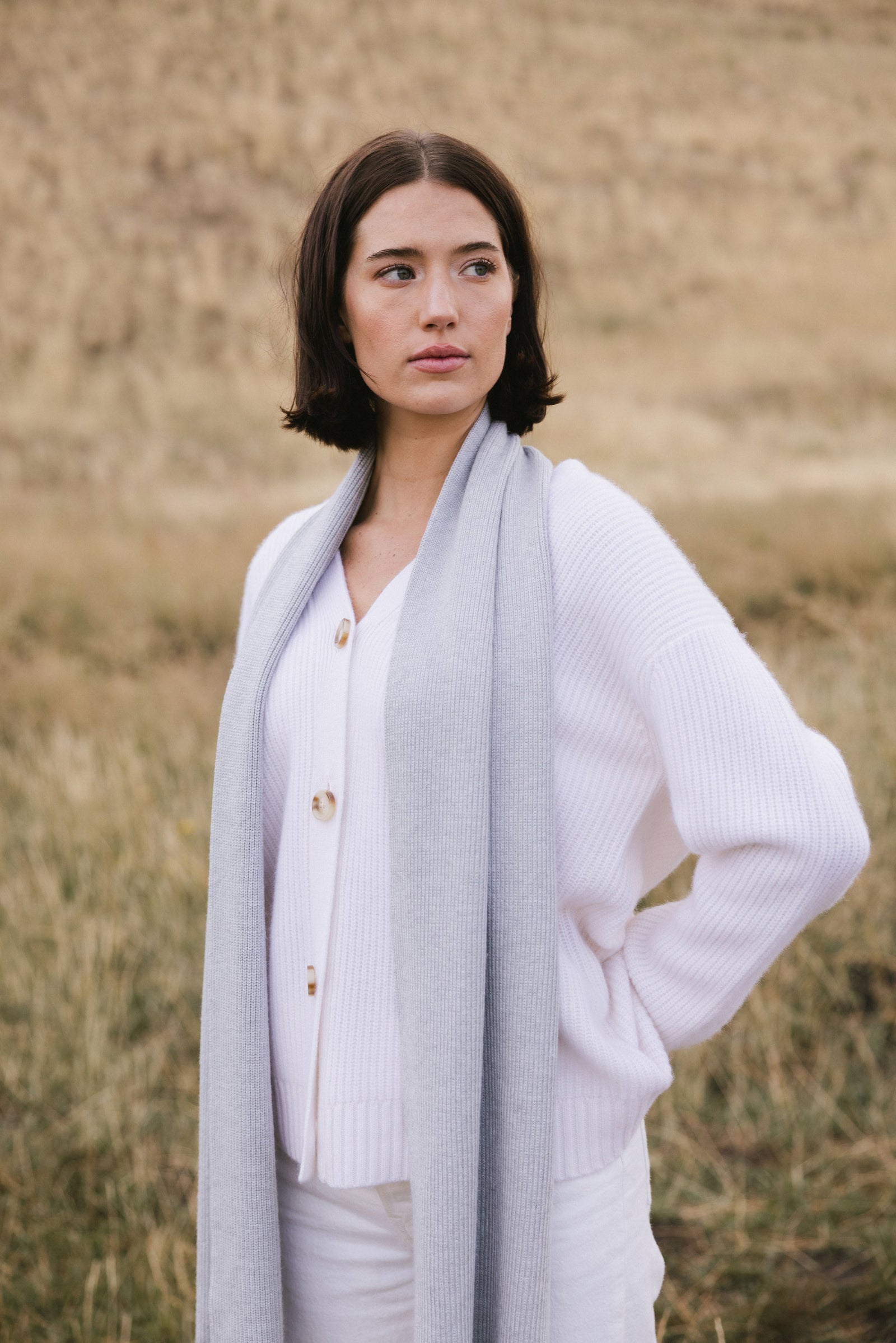 Woman wearing a light grey scarf, white sweater, and light-colored pants in a field