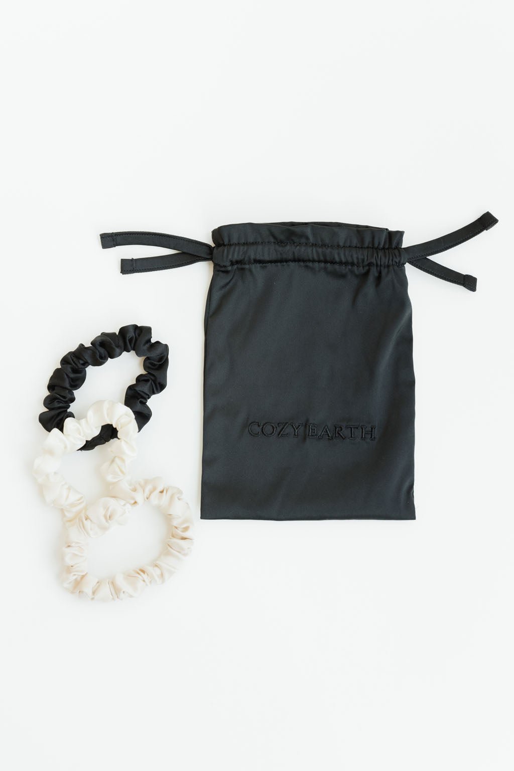 The image displays Cozy Earth's Silk Skinny Scrunchies in black, cream, and beige colors alongside a black drawstring bag embroidered with "COZY EARTH." The items are showcased on a white background.