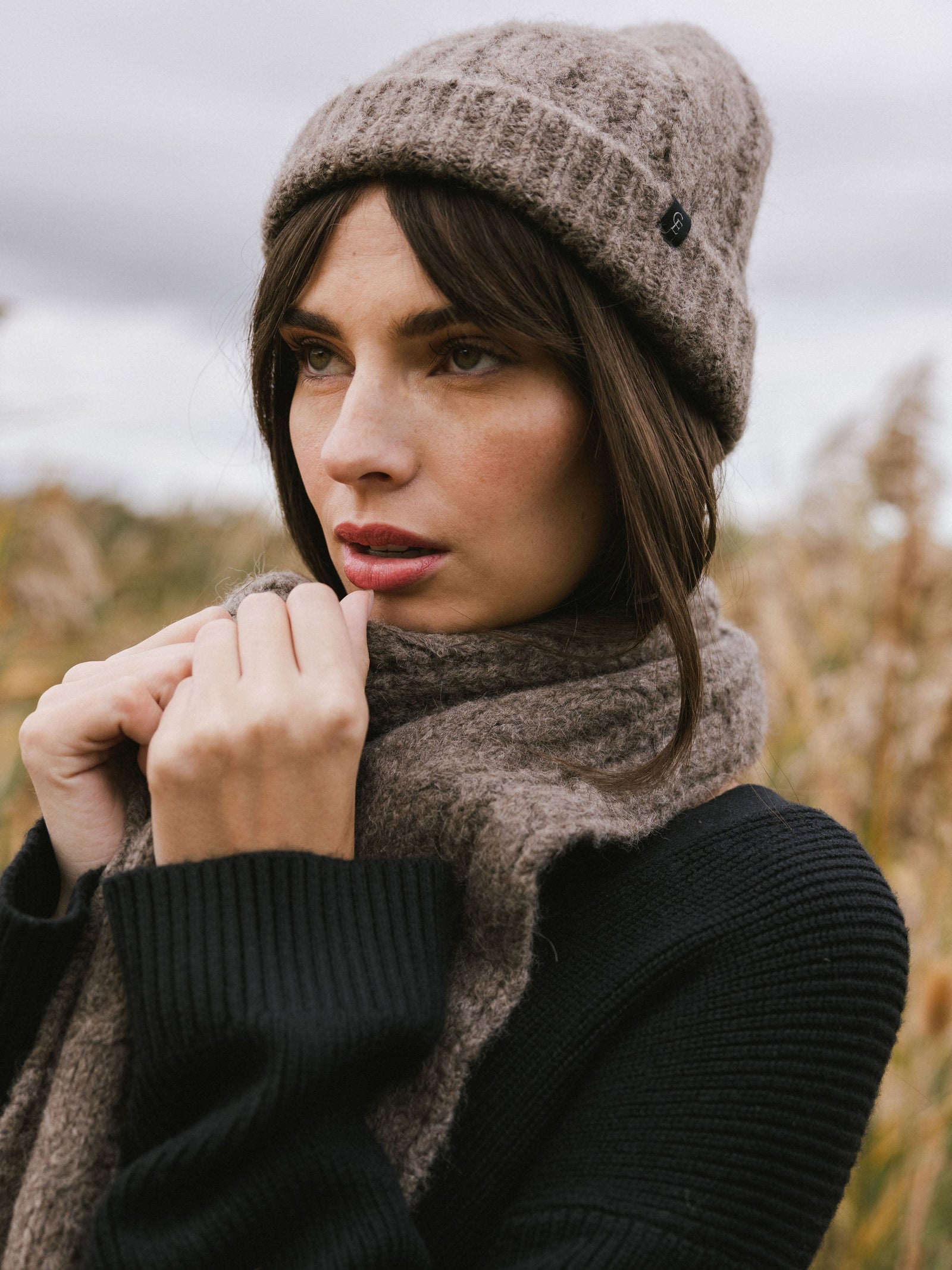 Woman wearing a gray Cozy Earth Cable Knit Beanie and scarf in a field of tall grass.