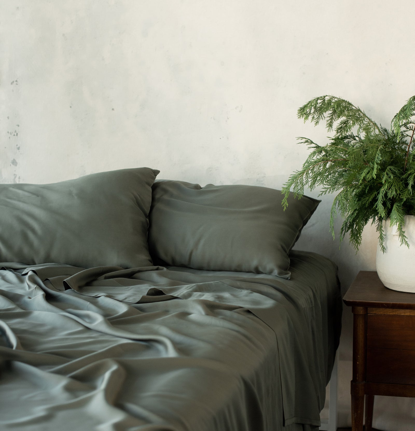 Unmade bed with olive bedding and a plant on the nightstand 