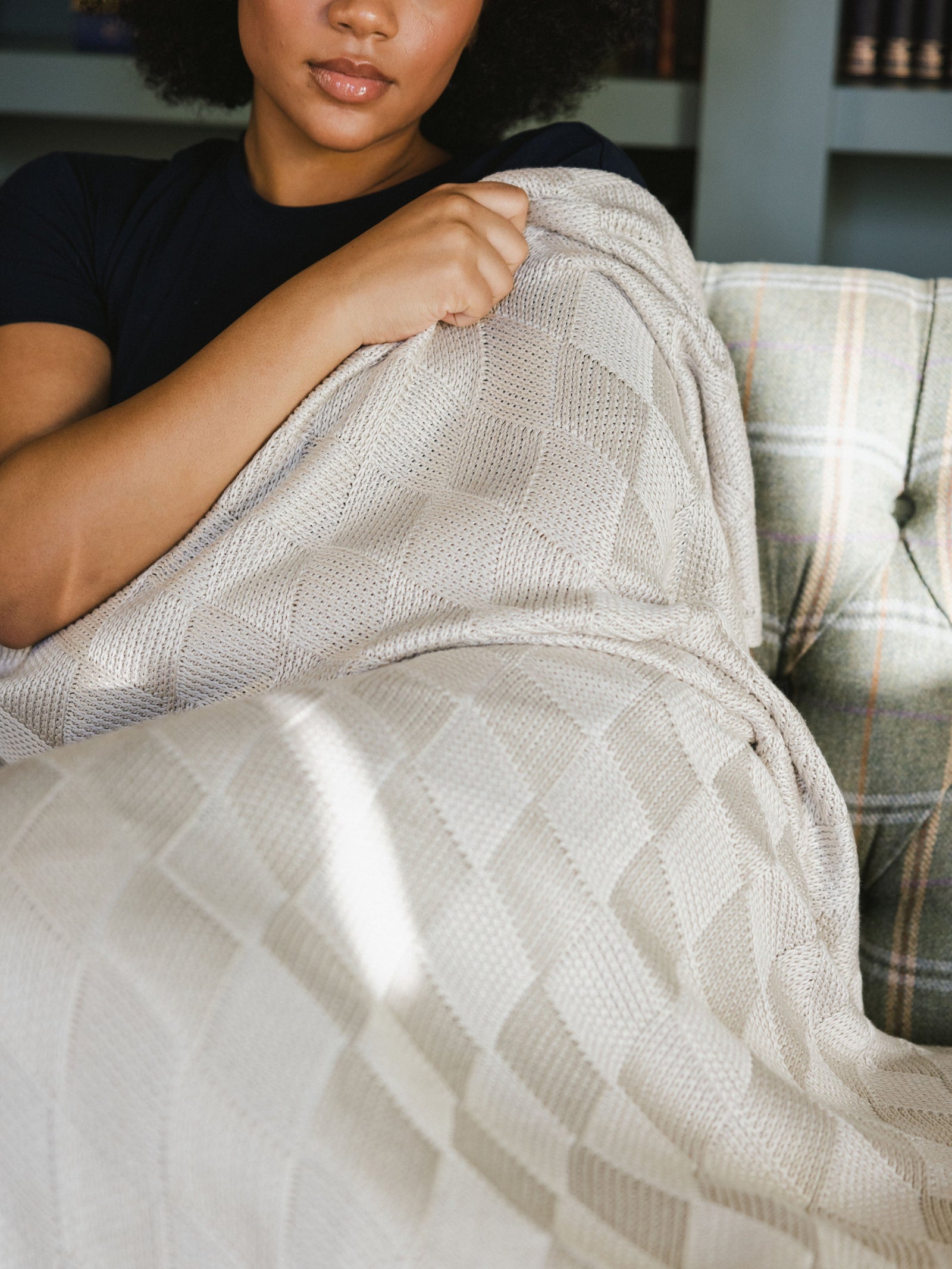 Beige Diamond Knit Blanket photographed in a home living room. A woman holds the blanket over her shoulder. 