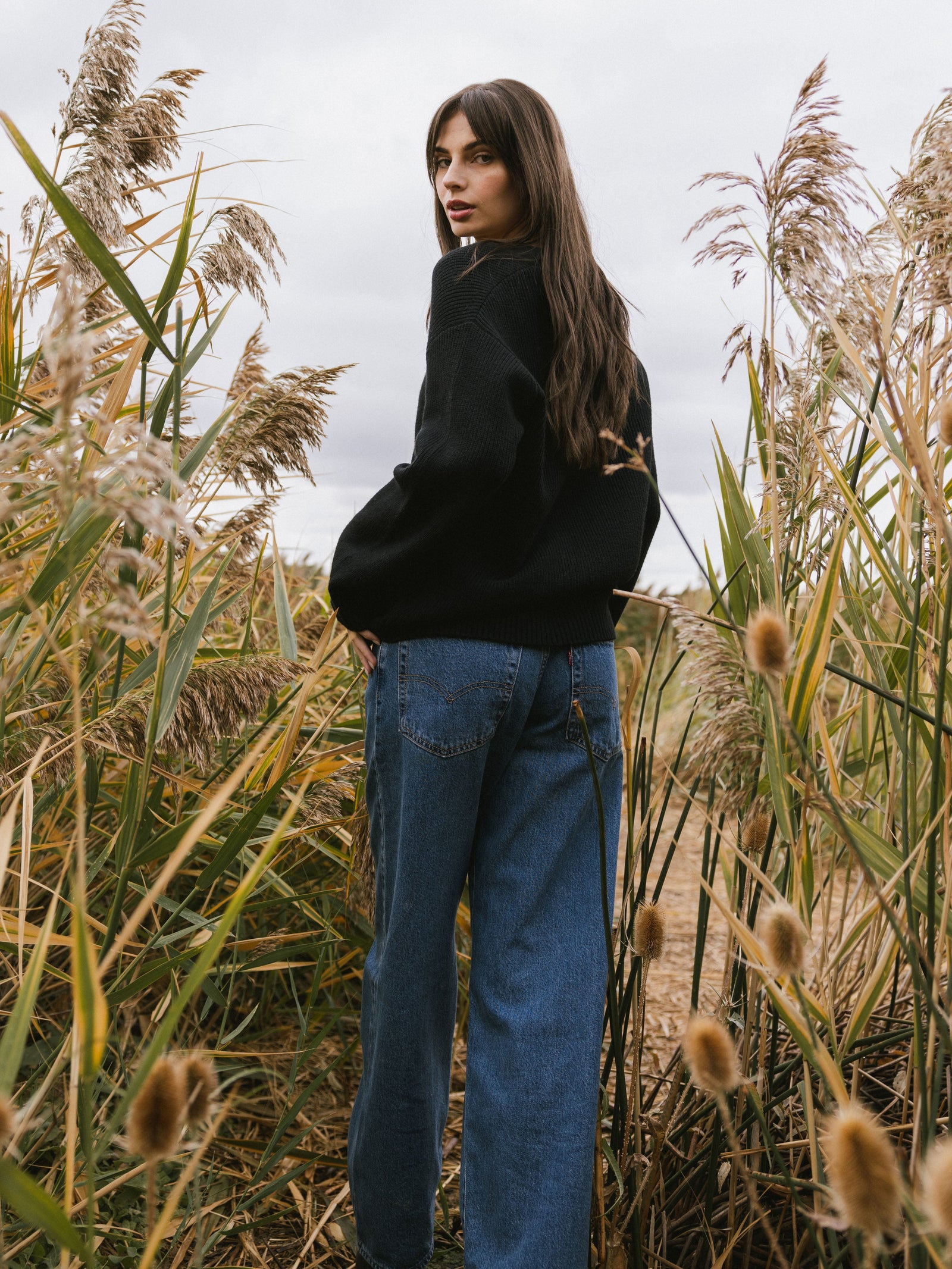 A woman stands surrounded by tall grass wearing Cozy Earth Classic Cardigan in Black. She is walking away from the camera with her face looking back