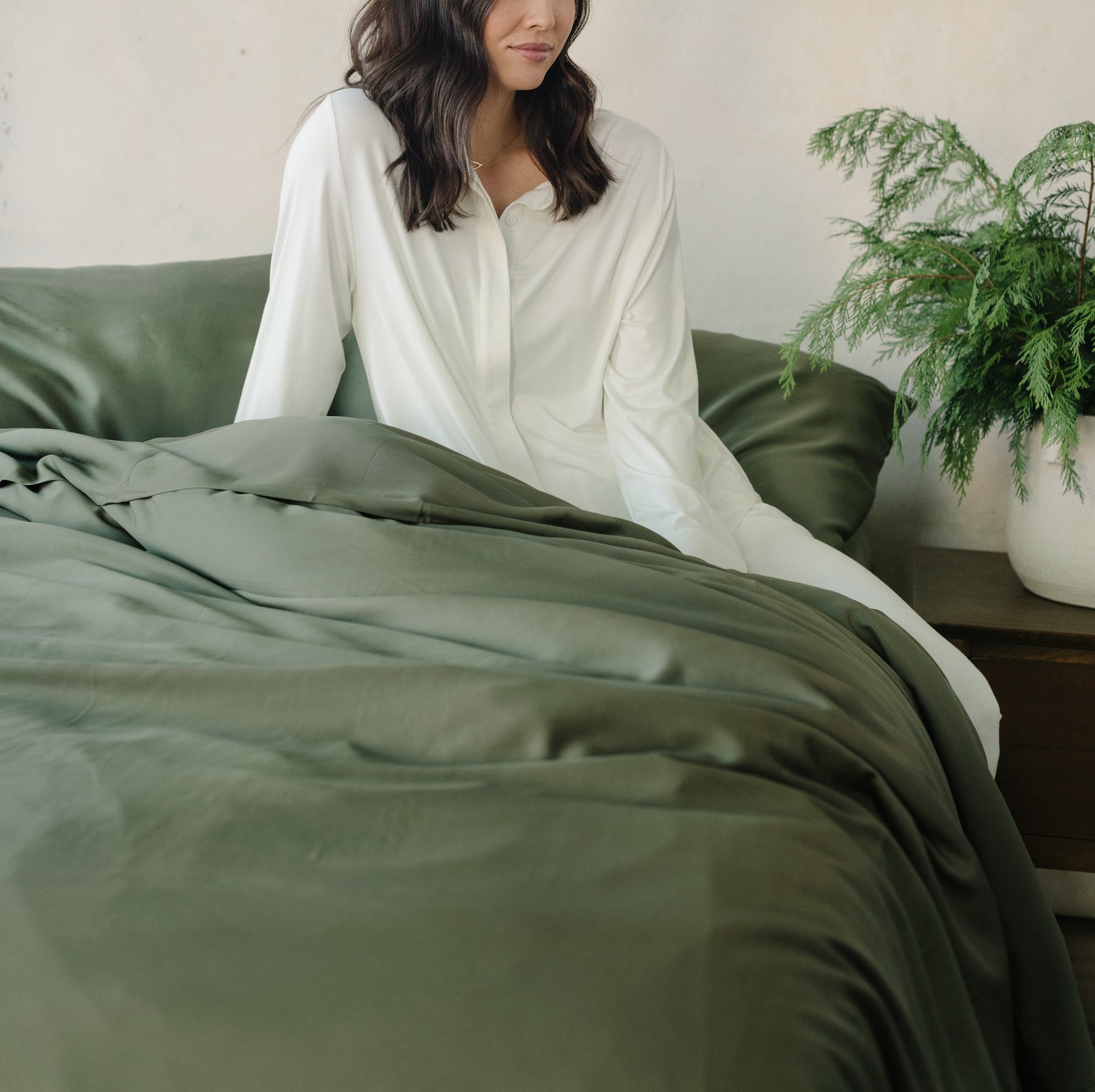Woman in white pajamas sitting on bed with olive bedding 