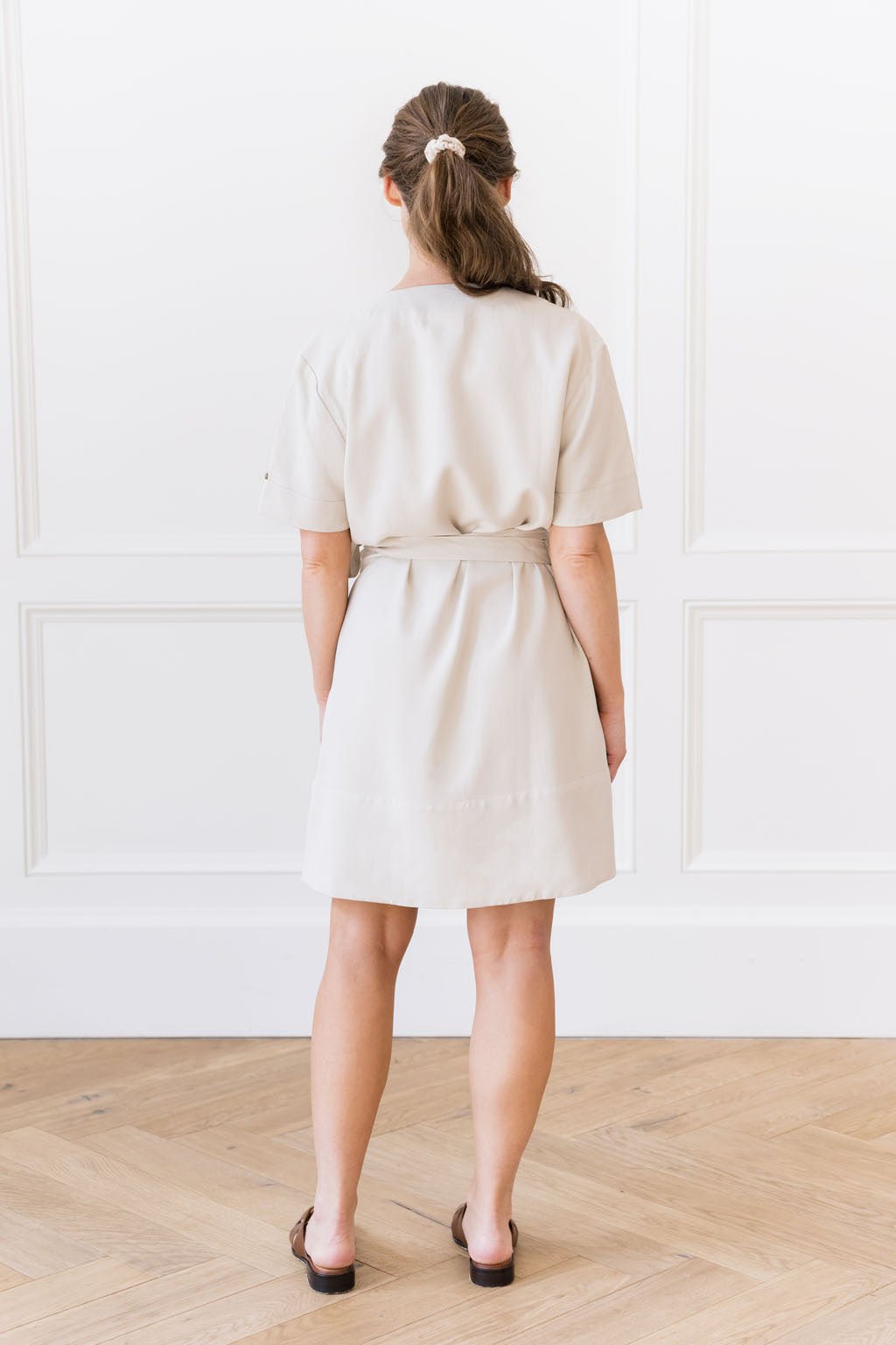 A woman stands with her back to the camera, wearing the Women's Cozy Earth Coastal Comfort Wrap Dress by Cozy Earth, which is short-sleeved and belted at the waist in a light beige color. Her dark hair is tied back in a ponytail with a white scrunchie. The background features a white wall with decorative paneling and a light wood floor.