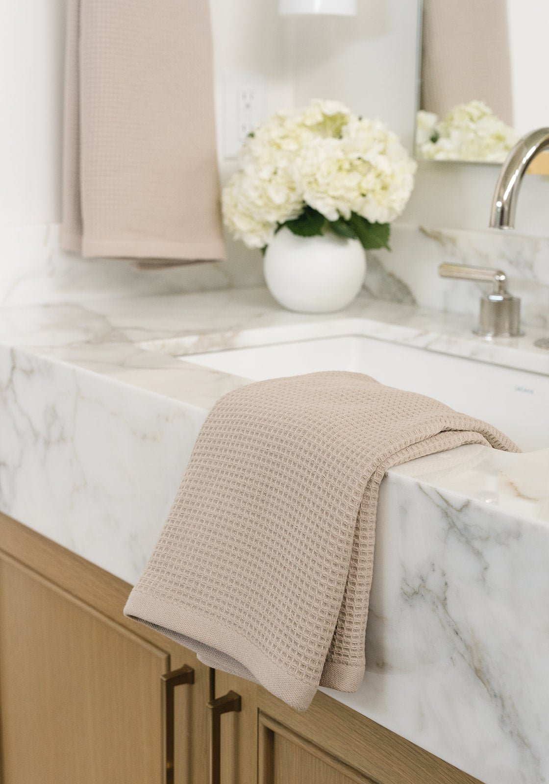 Sand Waffle Hand Towel resting folded on a white marble bathroom sink. The towel is folded so that different views of the towel can be seen. Another Hand Towel hangs from a towel ring in the background 
