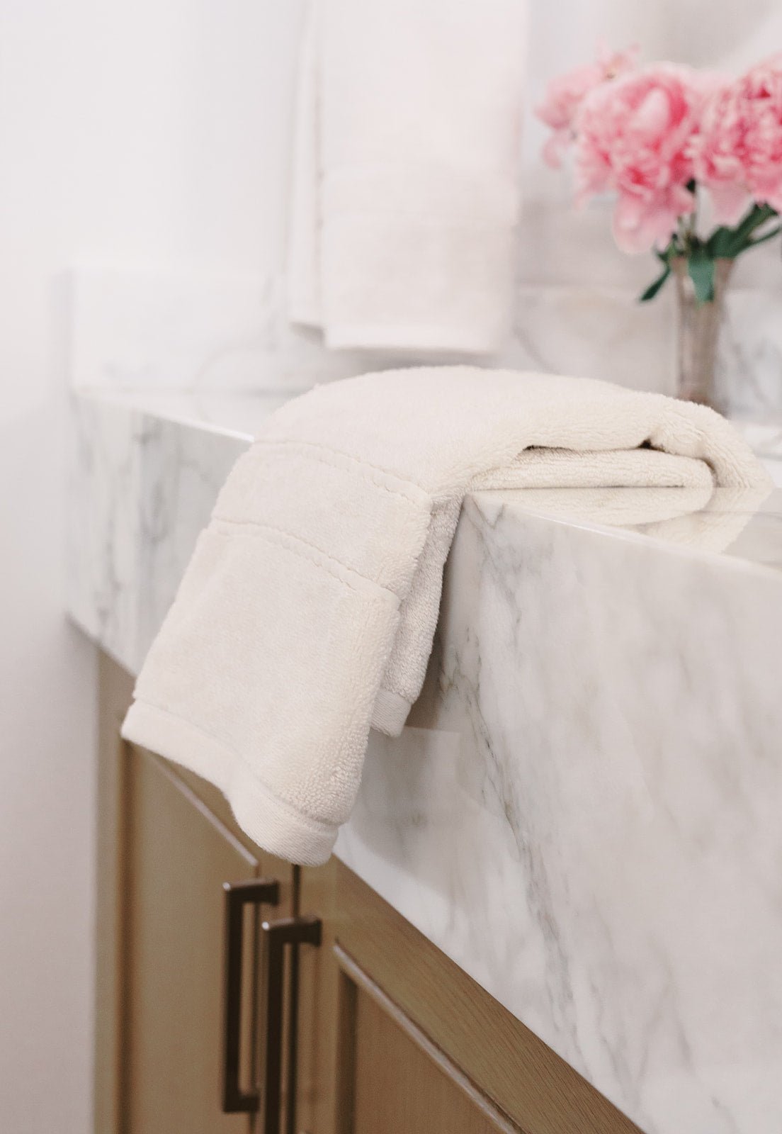 Premium Plush Hand Towel in the color Seashell. Photo of Seashell Premium Plush Hand Towel taken in a bathroom. One Premium Plush Hand Towel is hanging from a towel ring while the other is resting on the sink in the bathroom.