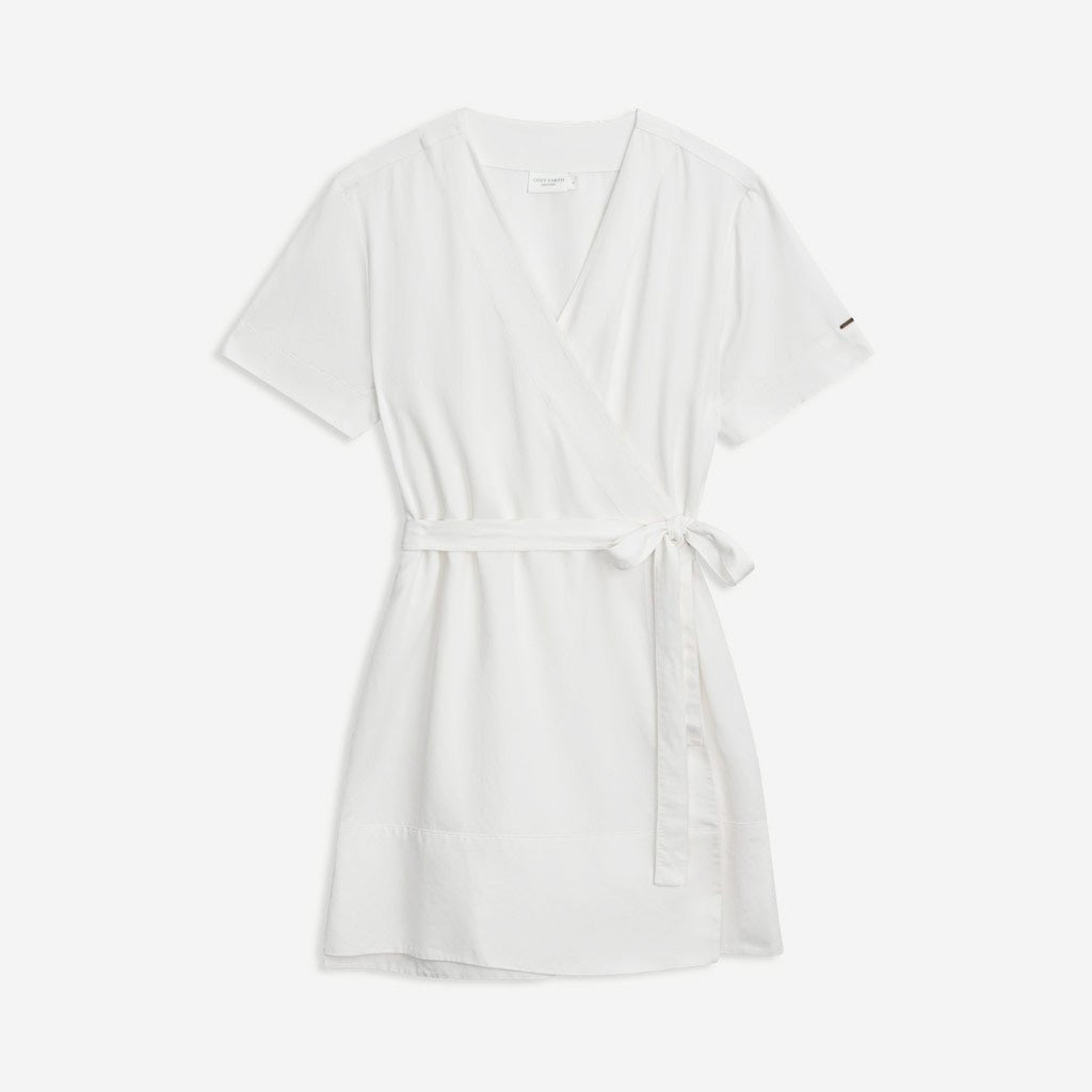 The Women's Cozy Earth Coastal Comfort Wrap Dress by Cozy Earth is a short, white wrap dress featuring short sleeves and a V-neckline. It is tied at the waist with a matching fabric belt and boasts a simple yet elegant design, making it ideal for casual or semi-formal occasions. The background is plain white.