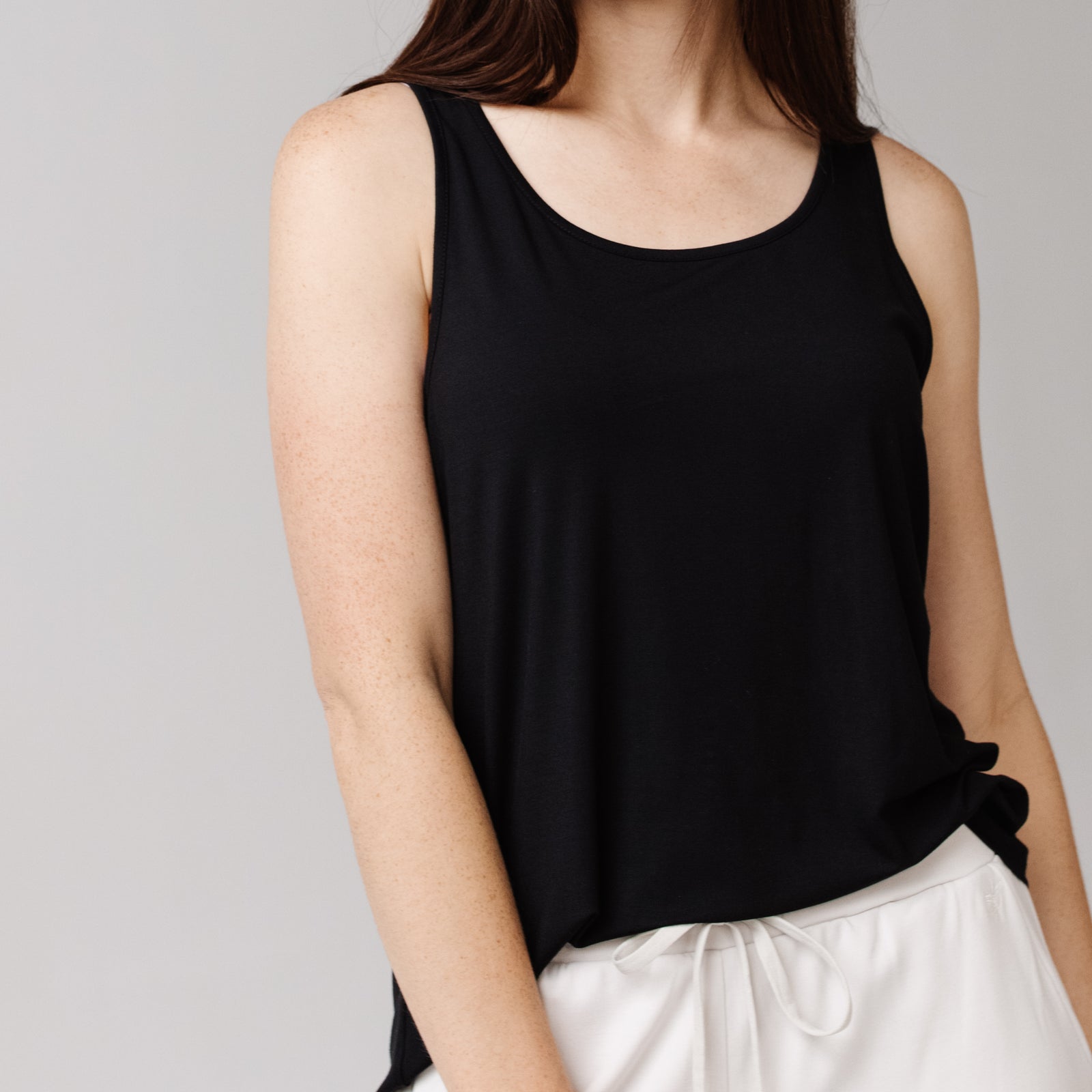 Woman in a black Cozy Earth Stretch Knit Tank and white shorts is shown from the neck down with a neutral background