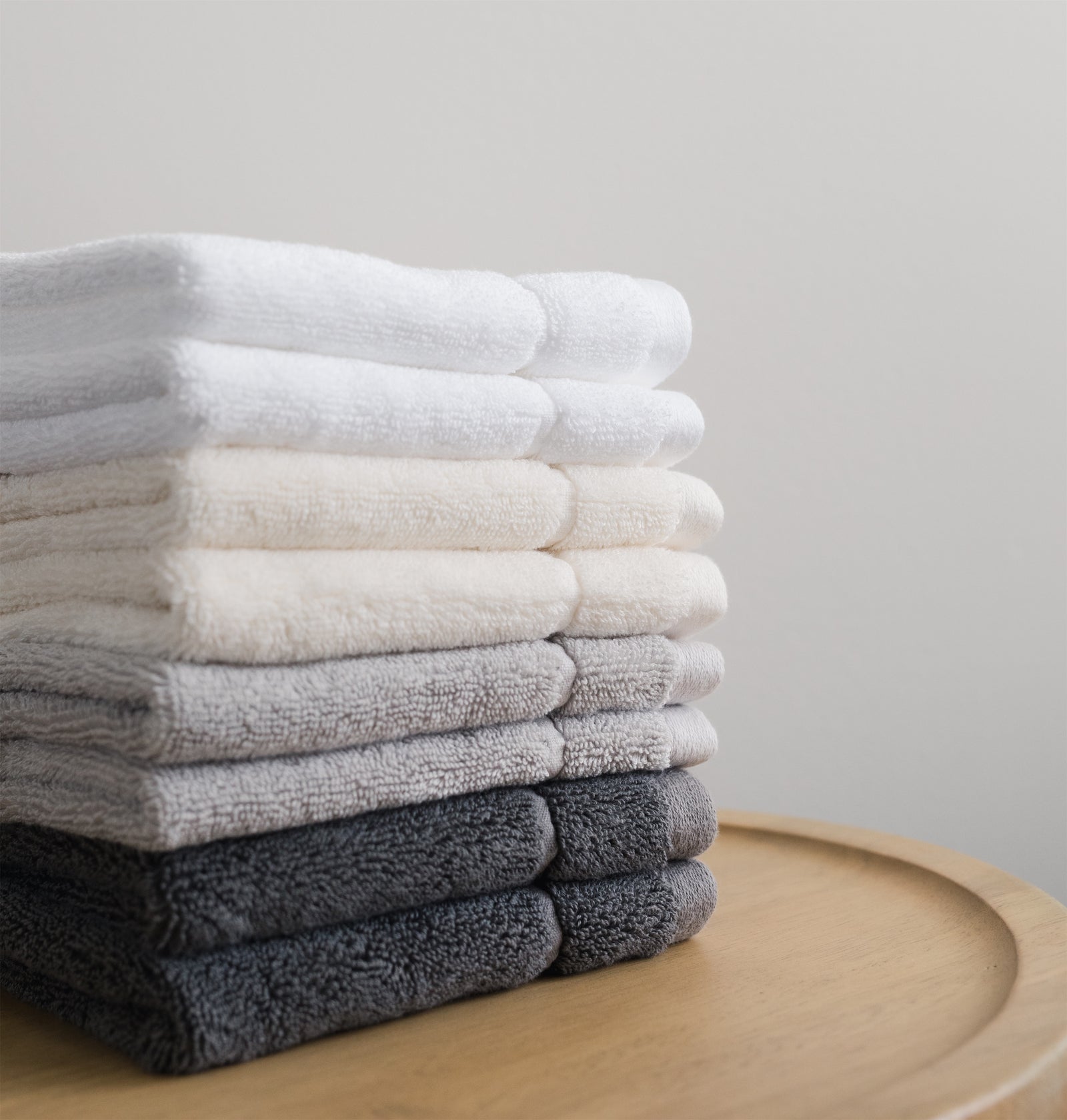 White, Light Grey, Charcoal, Creme Wash cloths stacked on top of each other. There is a white wall in the background. The towels are resting on a wooden stool.