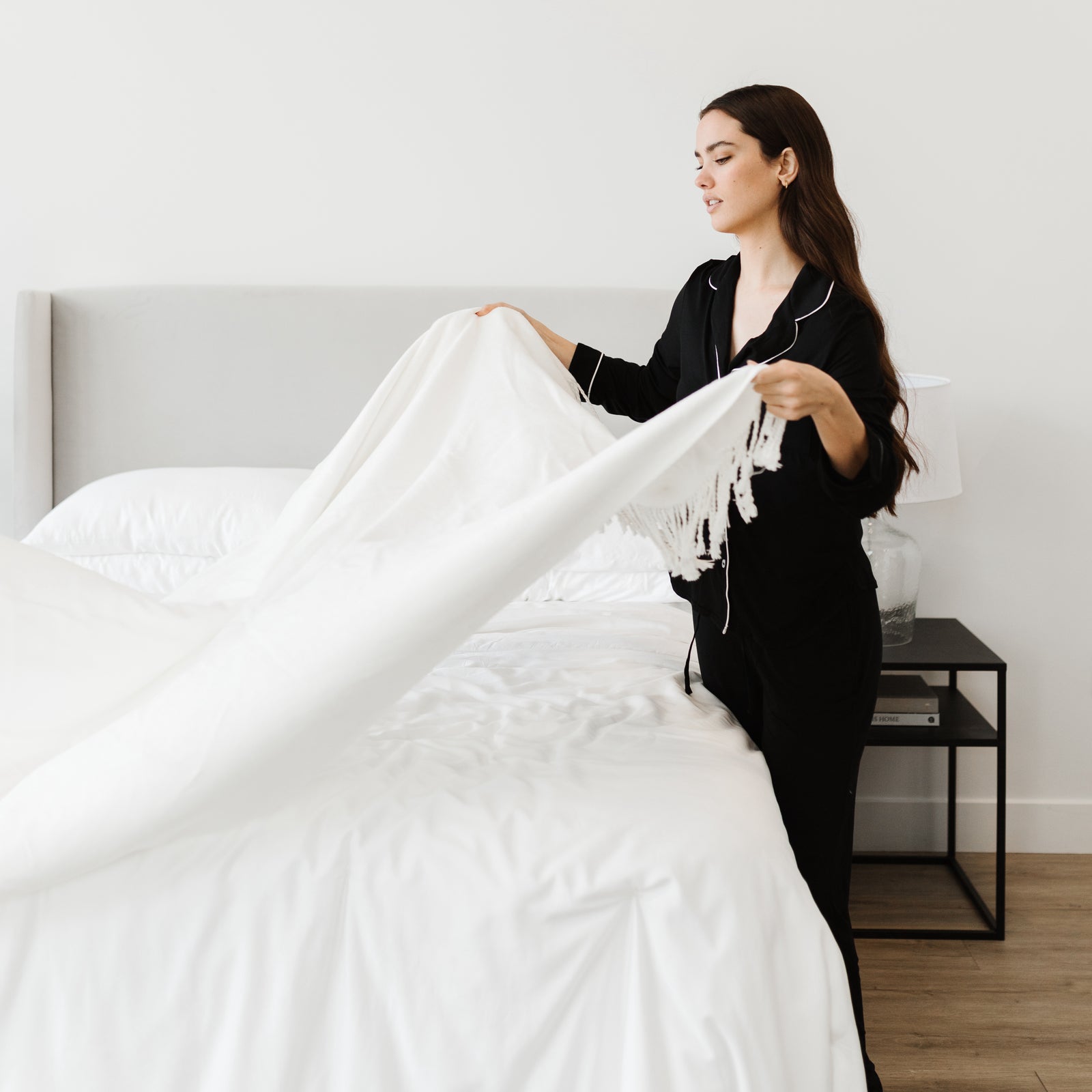 Woman in black pajamas laying throw blanket over white bed