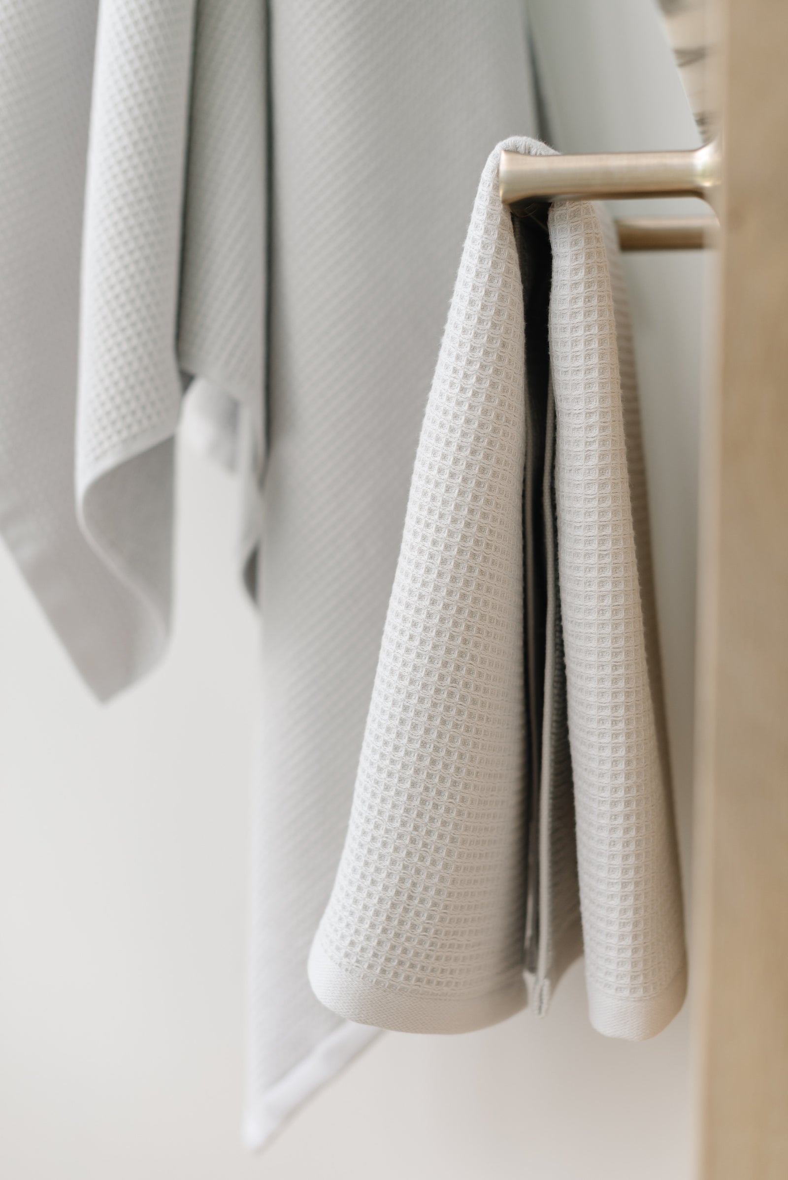 Light Grey Waffle Hang Towel handing from a gold towel bar.  The photo was taken at a white marble bathroom sink. A Waffle Bath Towel is seen hanging on the wall in the background. 