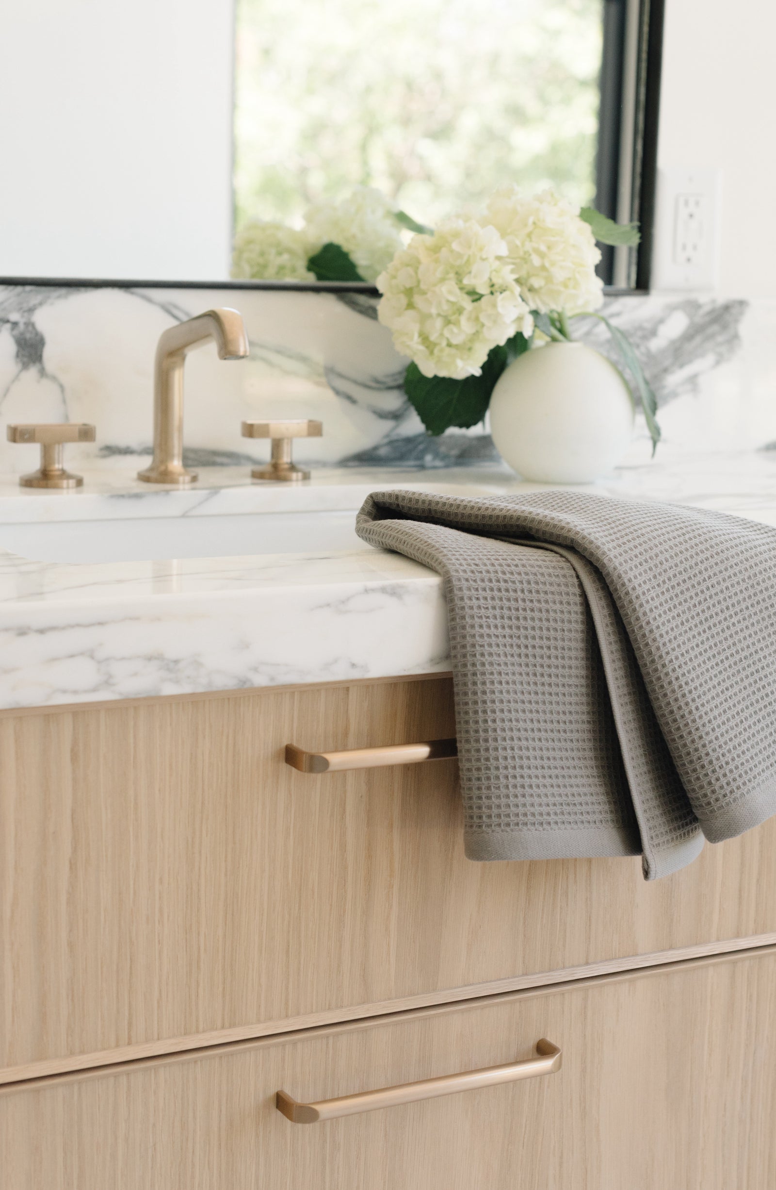 Charcoal Waffle Hand Towel resting folded on a white marble bathroom sink. The towel is folded so that different views of the towel can be seen. 