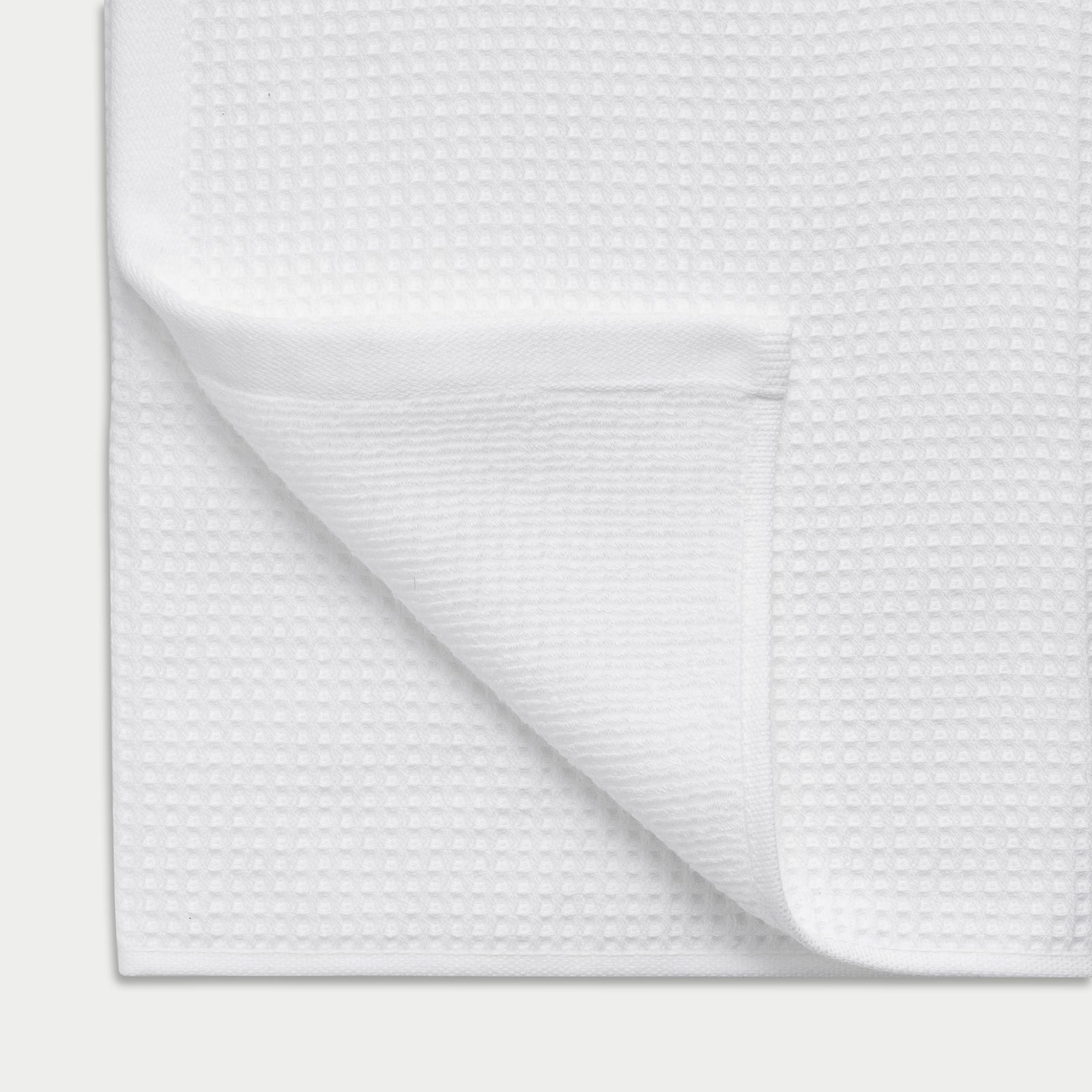 White Waffle Hand Towel resting flat on a white background. The corner is folded so that both sides of the towel can be seen. 